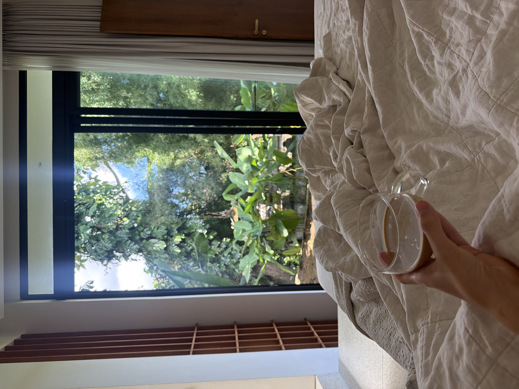 A medium shot captures a person's hand holding a glass cup containing a beige-colored liquid (likely a drink) while lying in bed. The bed has a wrinkled, light beige, quilted duvet. Outside the window, lush green foliage is visible. The window frame is dark, with horizontal bars. Below the window, there's a white surface with horizontal wooden slats. The image is set in an interior space in Mas, Indonesia. The scene appears to be capturing a calm morning moment.