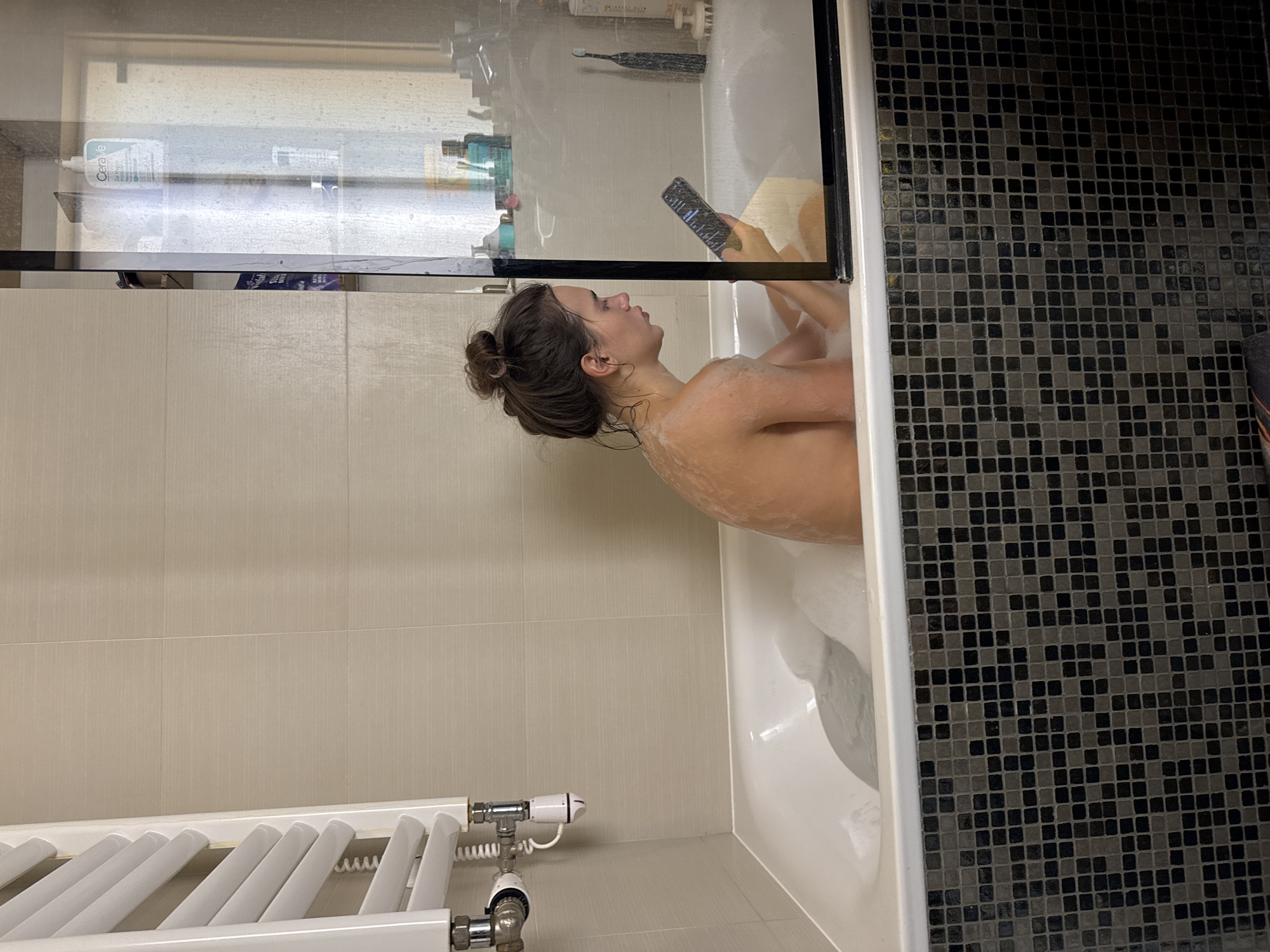 A person is in a bathtub, likely taking a bath with visible bubbles. The person is holding a smartphone in one hand, possibly interacting with it. The bathroom has tiled walls, a heated towel rack, and a glass partition around the tub. Shelves with various toiletries are visible behind the glass. The tiles are neutral and grey and the black tiles have a mosaic appearance.
Given the image characteristics and the absence of readily identifiable landmarks, pinpointing the exact location within Warsaw, Poland, is not possible.