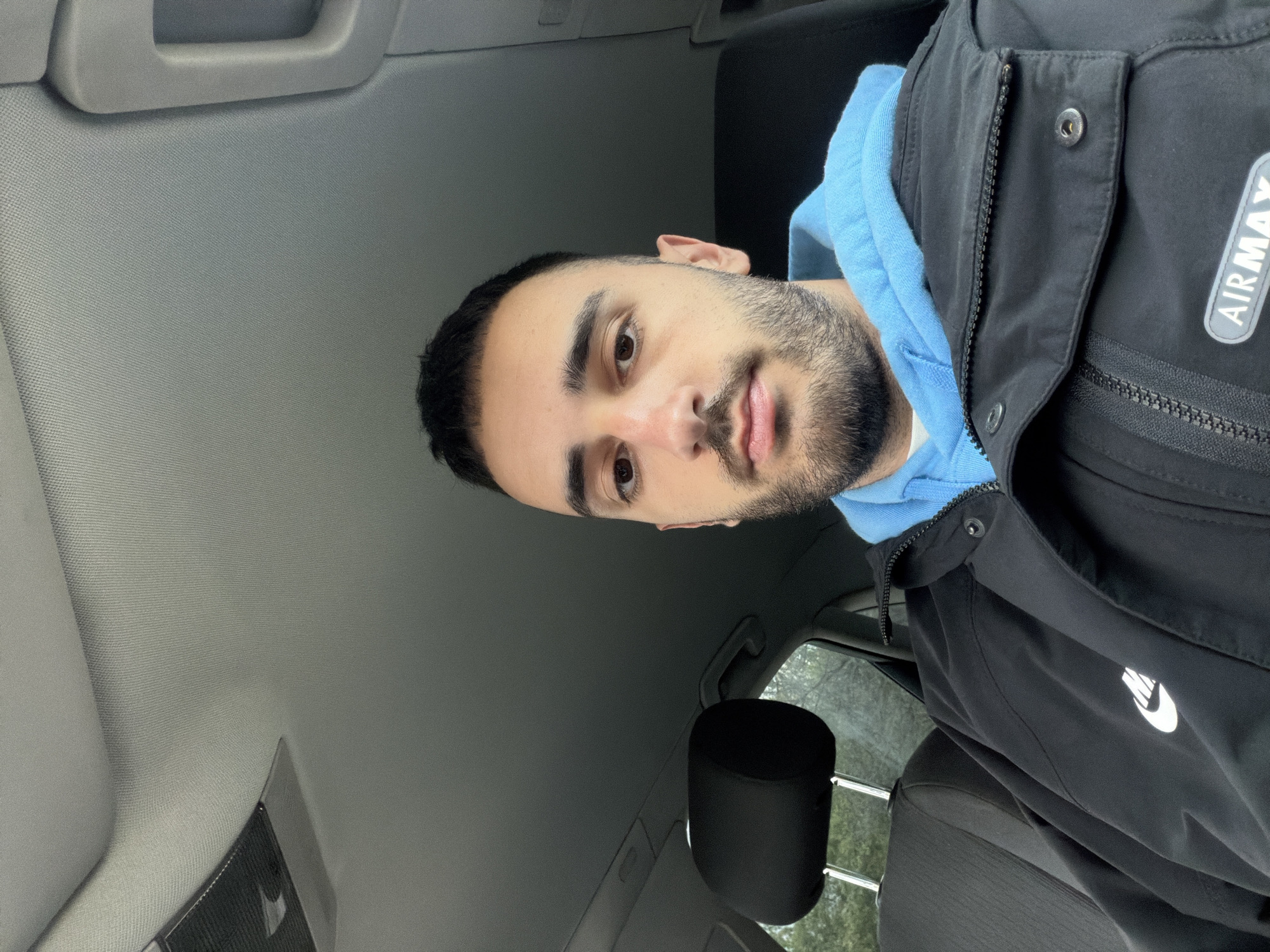 A man is sitting in the driver's seat of a car. He is wearing a light blue hoodie under a black jacket with "AIRMAX" visible on the right chest and a Nike swoosh on the left sleeve. The car's interior is gray. The photo appears to have been taken from a slightly angled, low perspective, with the camera looking upward.
There is not enough information in the image to determine the exact location in the city of Plovdiv, Bulgaria. The only visible context is the interior of a car.