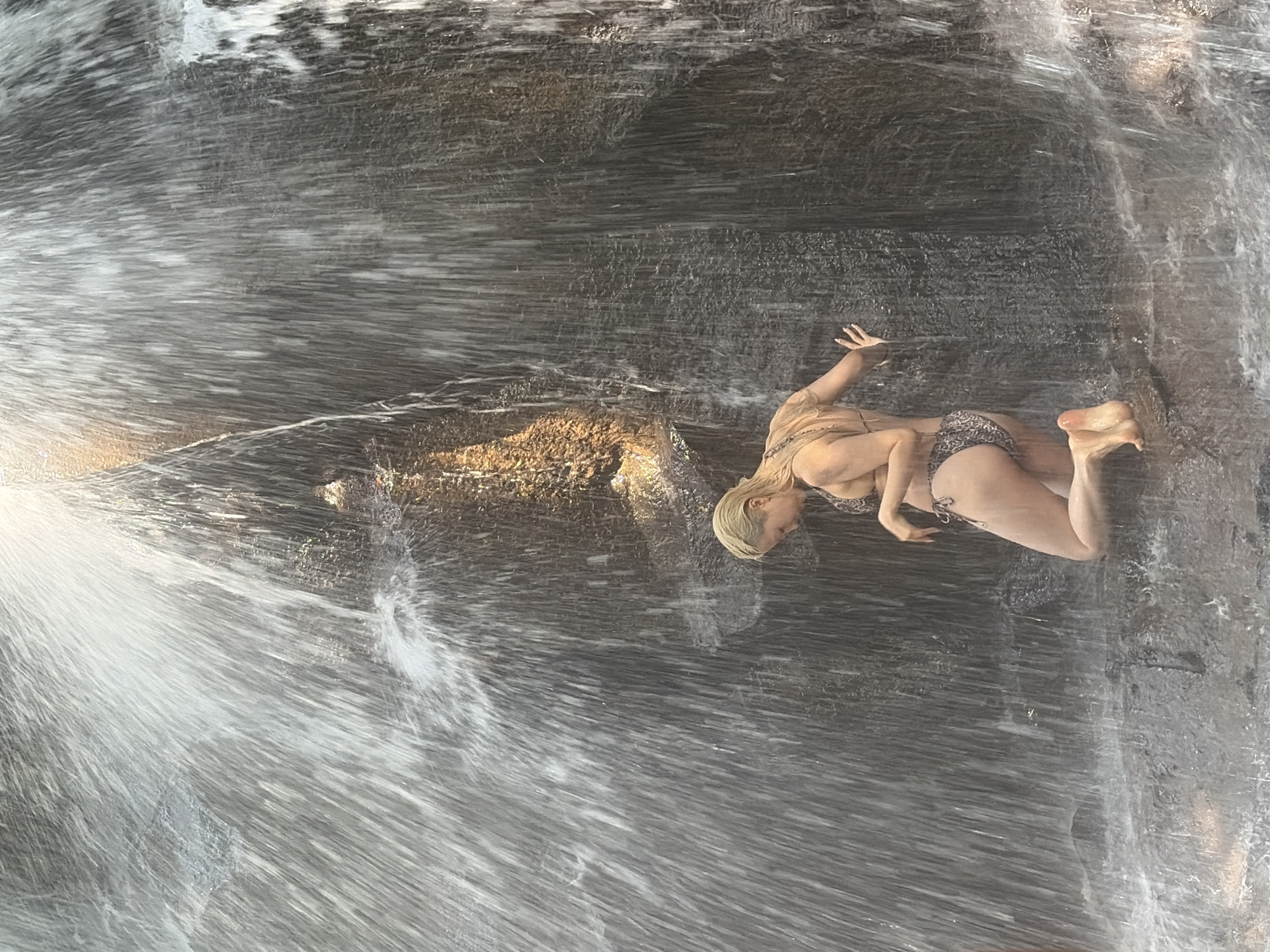 A woman in a leopard print bikini kneels on wet rocks in front of a waterfall, her back to the viewer. Water cascades down a dark, textured rock face, obscuring much of the background. The woman's blonde hair is wet, and she is looking down. Her left arm is extended, with her hand resting on the rock face. The location is believed to be in Gianyar, Indonesia, likely a natural waterfall setting.