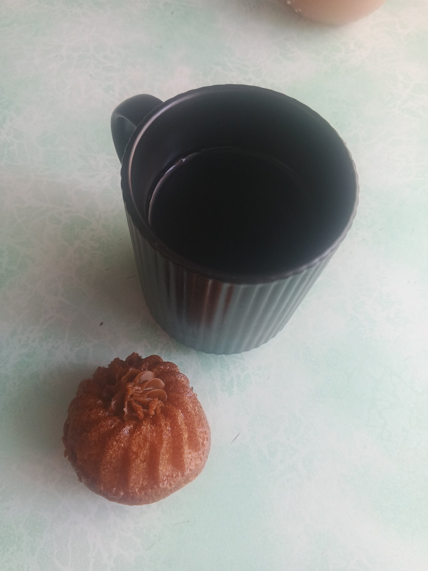 The image shows a black mug filled with dark liquid and a small, round cake with frosting on top, placed on a light-colored, patterned surface. The mug is ribbed and has a handle. The cake is golden brown and appears to be a bundt cake due to its fluted shape. The frosting on the cake is a light brown color and has a swirled pattern. The surface the items are on has a subtle, marble-like pattern in light green and white. The image is taken from a slightly elevated angle, looking down at the objects. There is no indication of the specific location within Biysk, Russia.