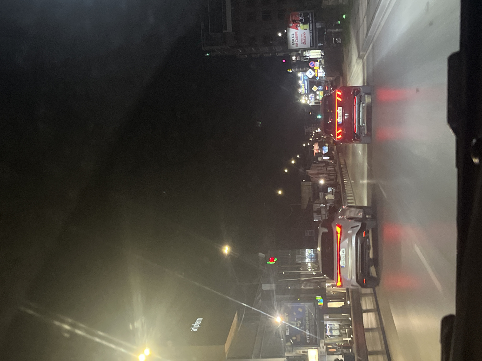 The image depicts a night scene in Sofia, Bulgaria, taken from the interior of a vehicle looking out towards the road ahead. Two cars are visible, a red one and a white one, both with their rear lights illuminated. Streetlights line the road, illuminating buildings and billboards. In the lower left, a building with "Atopharm" signage can be seen. Further up the road, there are other buildings and signs including ads for "Kасата на събира", Inbet, and Delano. The general environment is urban with street lighting and cars traveling on a road at night.
