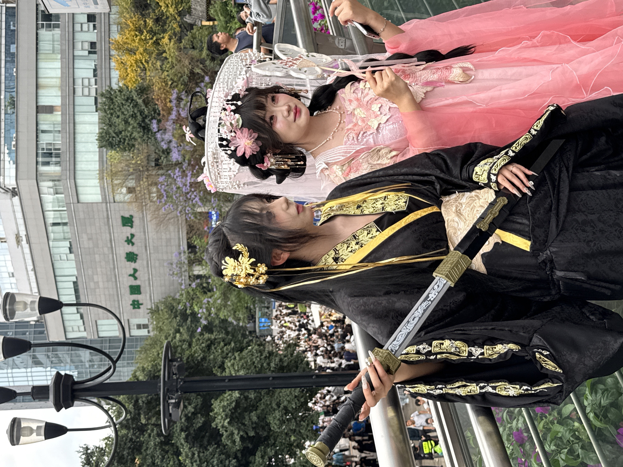Two women pose on a pedestrian overpass in Yuzhong District, China. One woman, lying down, wears a pink traditional dress with floral decorations and a white headdress; she holds a phone. The other woman, standing, wears a black traditional dress with gold trim and holds a decorative sword. Below them is a crowd of people. In the background are trees, a street lamp, and a building with Chinese characters on it.