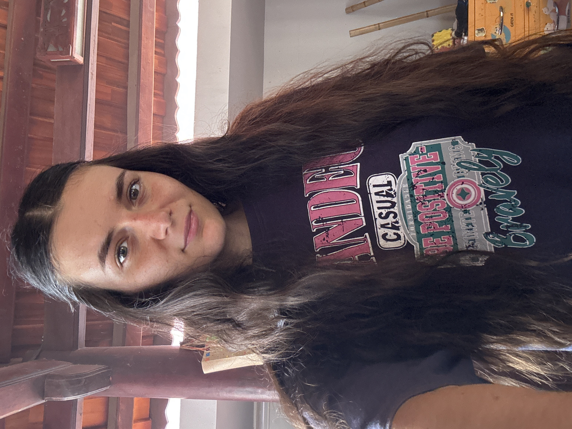 A person with long brown hair is facing the camera indoors. The person is wearing a dark shirt with a graphic design that includes the words "ANDER," "CASUAL," "FORMING," "BE POSITIVE," and "Bravely." The background shows part of a wooden ceiling structure and a light-colored wall or window. The location is Mũi Né, Vietnam.