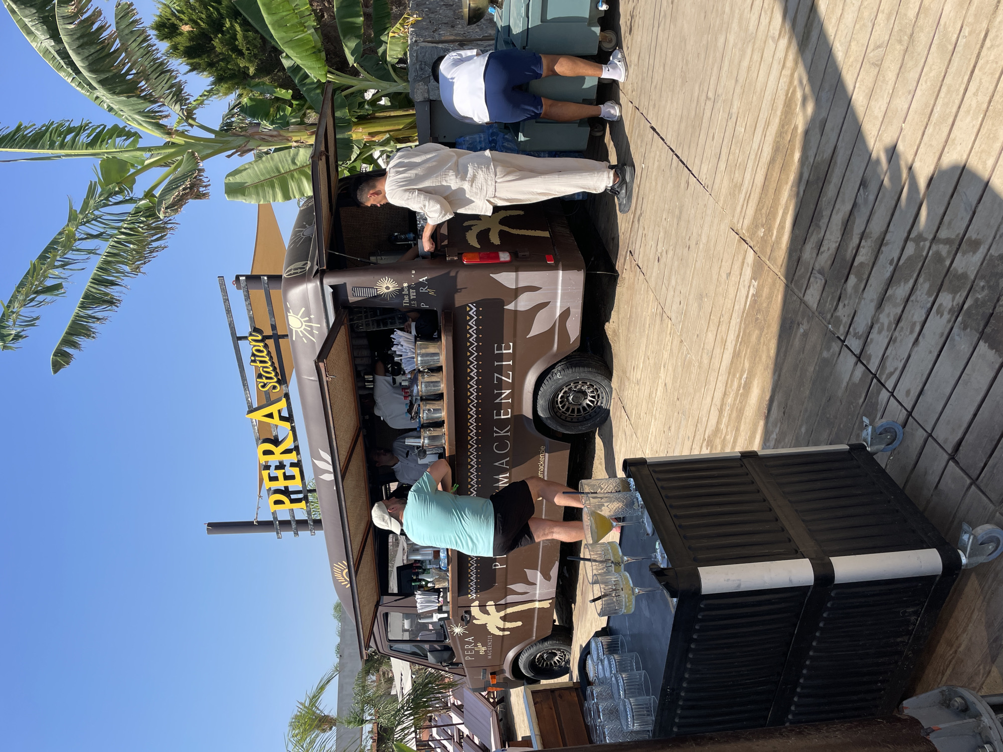 The image depicts a mobile bar, labeled "PERA Station," situated outdoors on a wooden deck, likely in Trikomo, Northern Cyprus. A prominent yellow sign with black lettering reading "PERA Station" is mounted above the bar. The bar itself is a brown van with an open service window. Inside, several individuals appear to be working, with one in a white uniform handing something to another person.
Outside the van, two men are visible. One man, wearing a light blue t-shirt and black shorts, is leaning on the service counter. Another man, dressed in a white linen outfit, is standing with his back to the camera, facing the bar. A third man, wearing a white t-shirt and blue shorts, is partially visible on the right side of the frame, appearing to reach into a large green cooler.
In the foreground, a black mobile cart holds several glasses, some filled with drinks, on its top surface. The environment is sunny with a clear blue sky. Lush green palm trees and other foliage are visible in the background, suggesting a tropical or Mediterranean setting. The wooden deck on which the bar is placed suggests a beachside or poolside location.