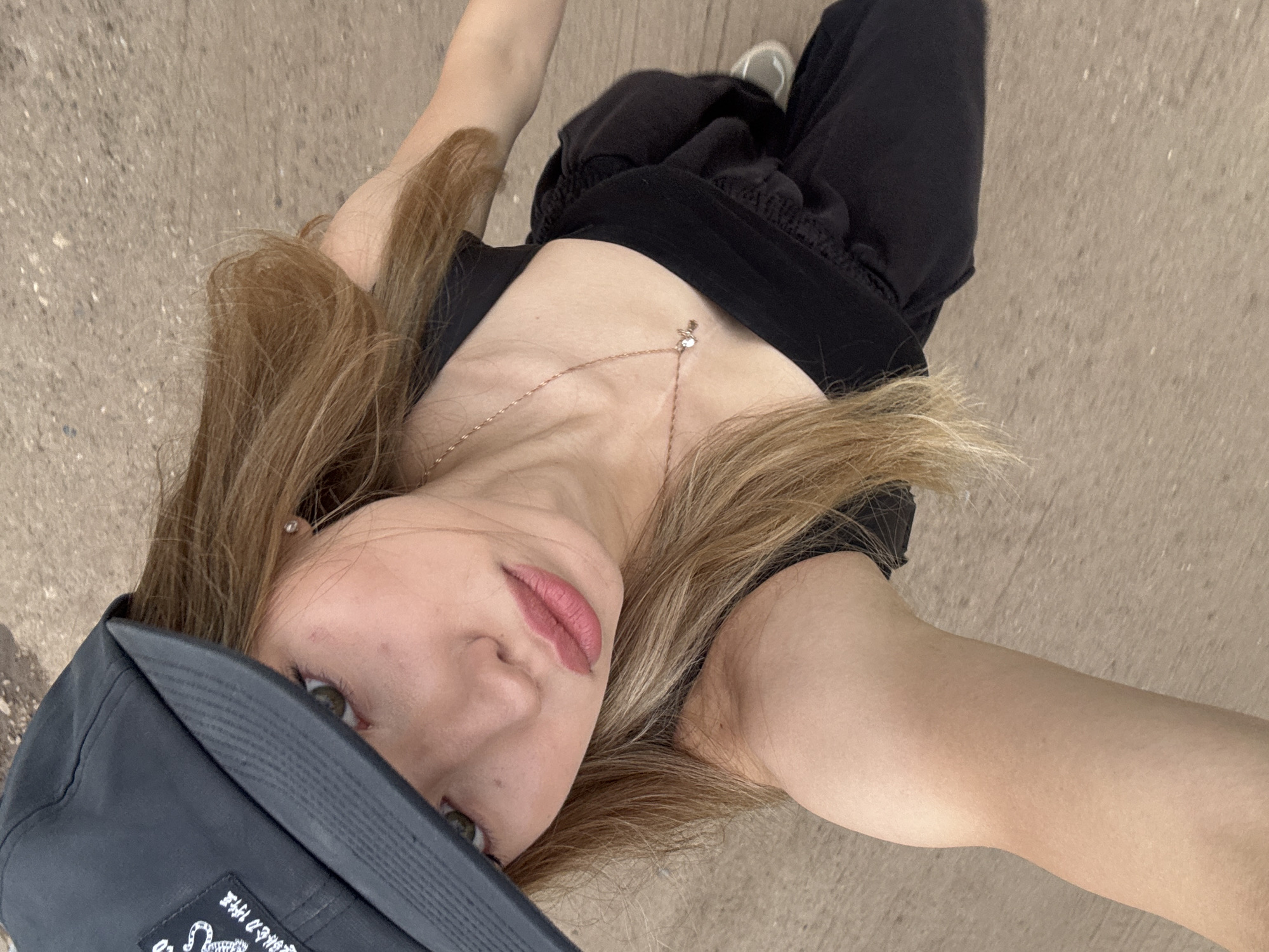 The image shows a young person wearing a black baseball cap and black clothing, including pants and a top. They have long, blonde hair and are wearing a necklace with a small pendant. The person is looking at the camera with their eyes visible and their lips slightly parted. The background appears to be a paved surface, possibly a sidewalk or road. The image is taken from a high angle, looking down at the person. There is some visible text on the baseball cap, but it is mostly obscured by the angle of the image. The lighting suggests it is daytime, and the weather appears to be clear. The overall mood of the image is casual and relaxed.