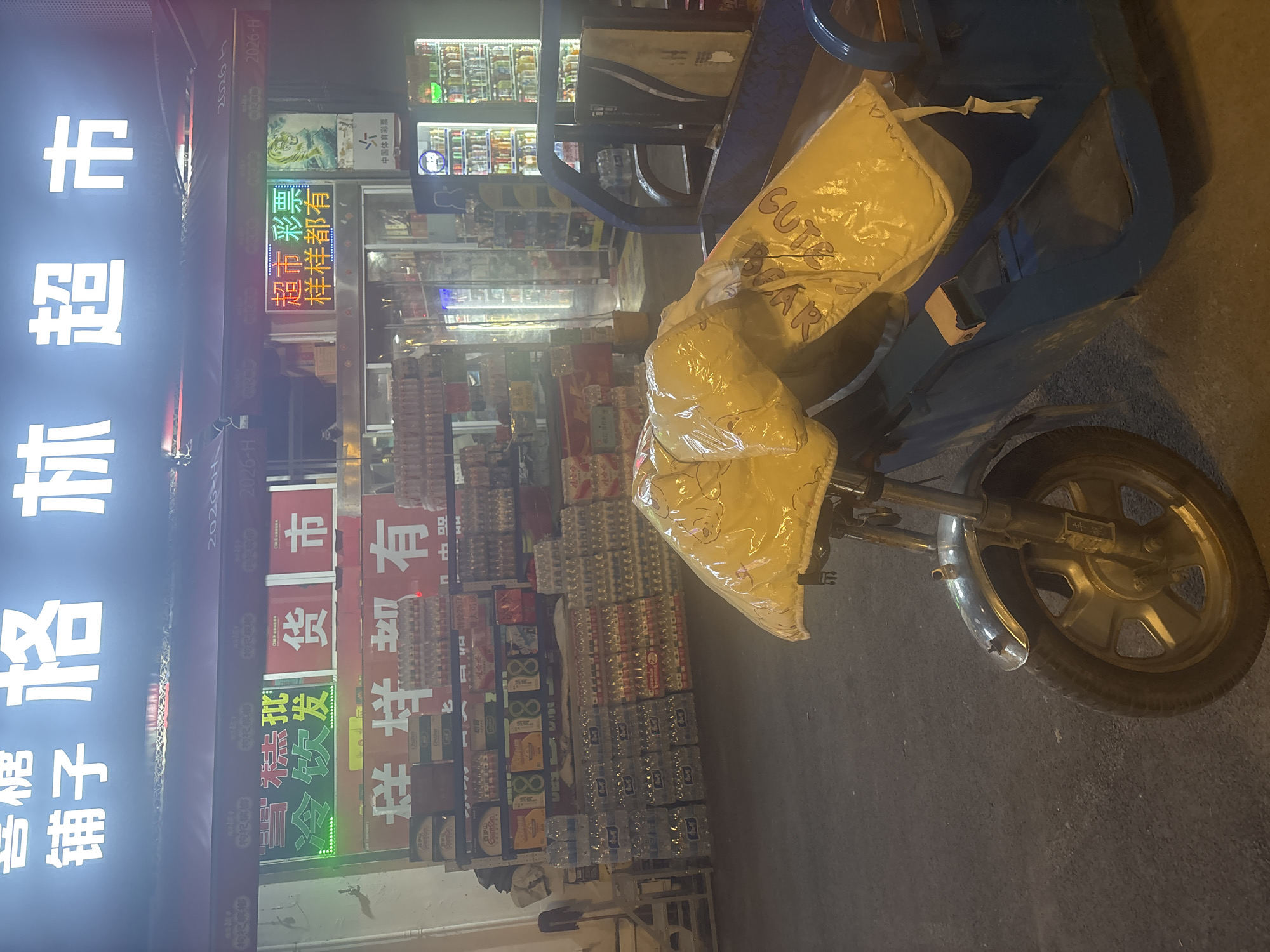 The image shows a small supermarket in Suzhou Industrial Park, China, at night. The supermarket's name, "格林超市" (Green Supermarket), is displayed in large, illuminated characters on its facade. Below this, a sign advertises "生鲜批发" (Fresh Produce Wholesale) and "冷饮发货" (Cold Drink Delivery). Further to the right, another neon sign states "超市彩票 样样都有" (Supermarket Lottery, Everything Available).
In front of the supermarket, a blue motorized tricycle is parked. The tricycle's cargo area is covered with two large yellow bags, one of which has a cartoon bear print. A large stack of bottled water is visible to the left of the tricycle, and rows of shelves stocked with various goods, including drinks and packaged foods, are visible inside the supermarket through its open entrance. The street in front of the supermarket is paved.