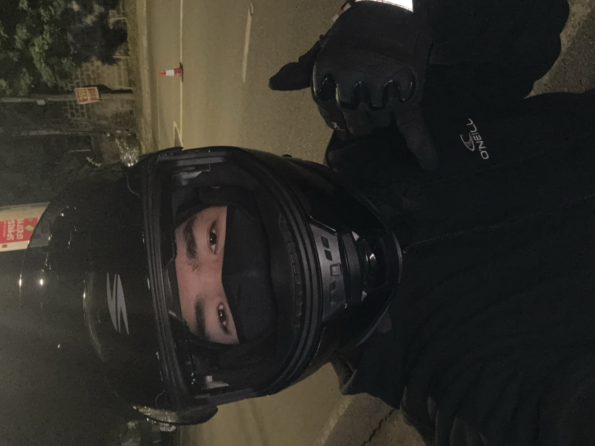 A person wearing a black motorcycle helmet, black face mask, and a black hoodie is in the foreground of the image. The person is giving a thumbs-up gesture with their right hand, which is covered by a black glove. The background shows a street at night with streetlights and some trees. There is a traffic cone and a brick wall with a sign on it. The location is identified as Davao City, Philippines.
