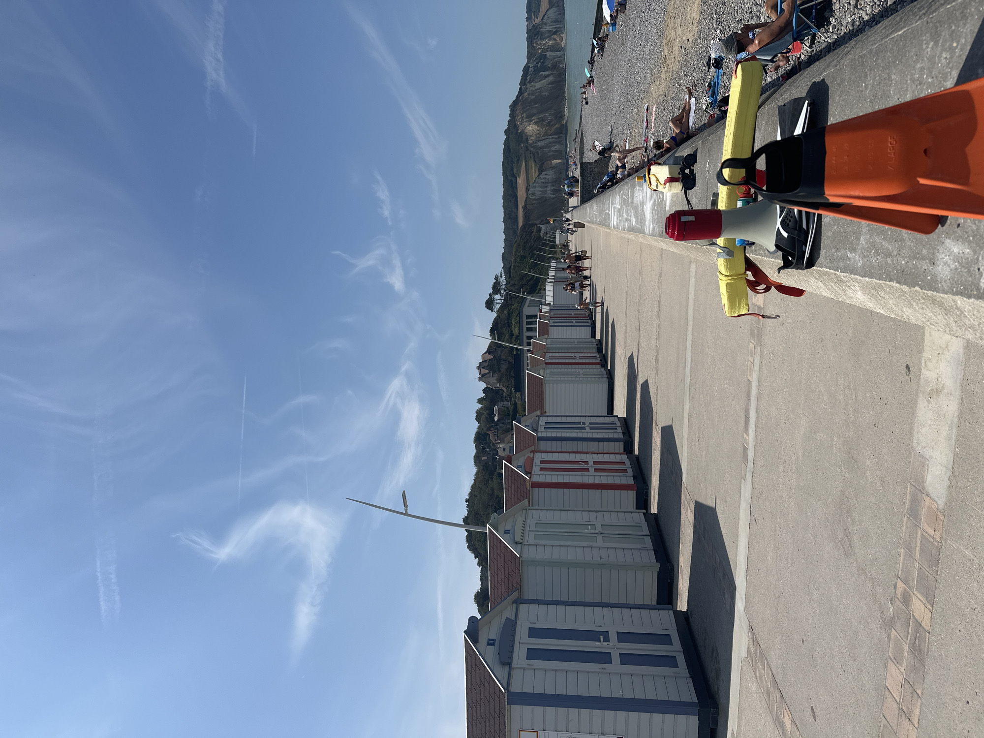 This image depicts a sunny day at the beach in Hautot-sur-Mer, France. In the foreground, a concrete barrier lines the edge of a paved walkway. Resting on the barrier are a pair of orange swim fins, a black and white towel, and a megaphone with a red top. Behind these items, a yellow flotation device with a red strap is visible.
The walkway stretches into the distance, lined with a row of small, white beach huts with various colored doors (blue, red, and light green). The huts have dark brown roofs. People are seen walking along the path, and further down, others are sunbathing on the pebble beach.
The beach itself is covered in grey pebbles and extends to the sea. In the background, a dramatic chalk cliff rises behind the beach, covered in green vegetation and dotted with buildings. The sky is a clear blue with wispy white clouds. The overall scene conveys a sense of a vibrant, recreational seaside environment.