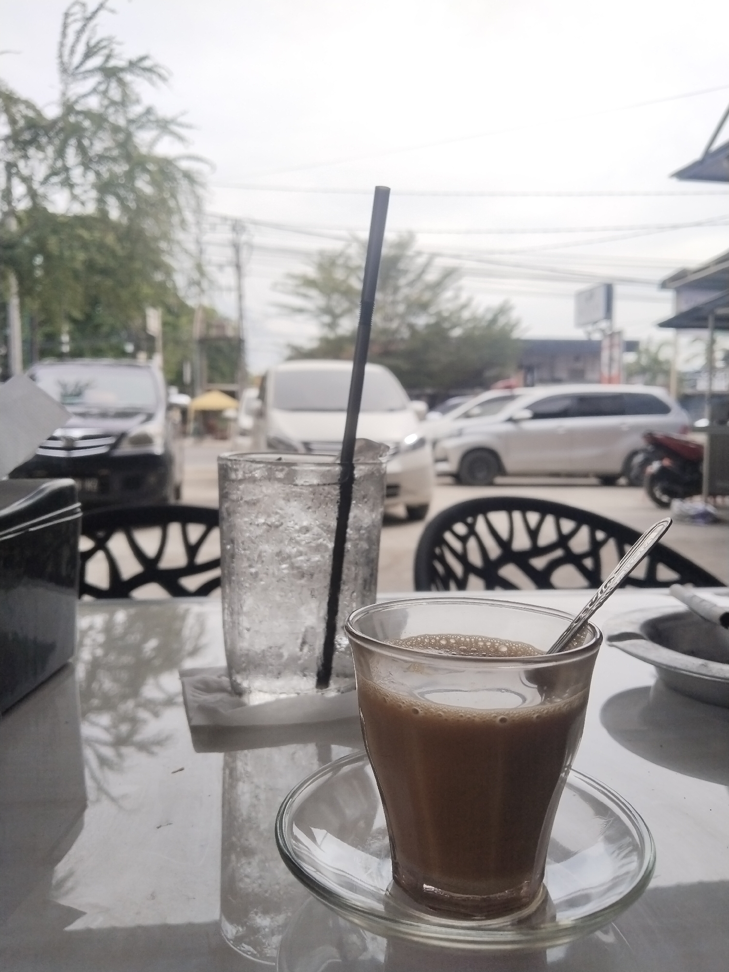 A close-up shot taken from a table in Lhokseumawe, Indonesia. On the table are a glass of iced water with a black straw, and a glass of coffee with a spoon inside, both on clear glass saucers. The table is white, and the background shows a street with cars, buildings, trees, and a motorcycle. Black chairs surround the table.