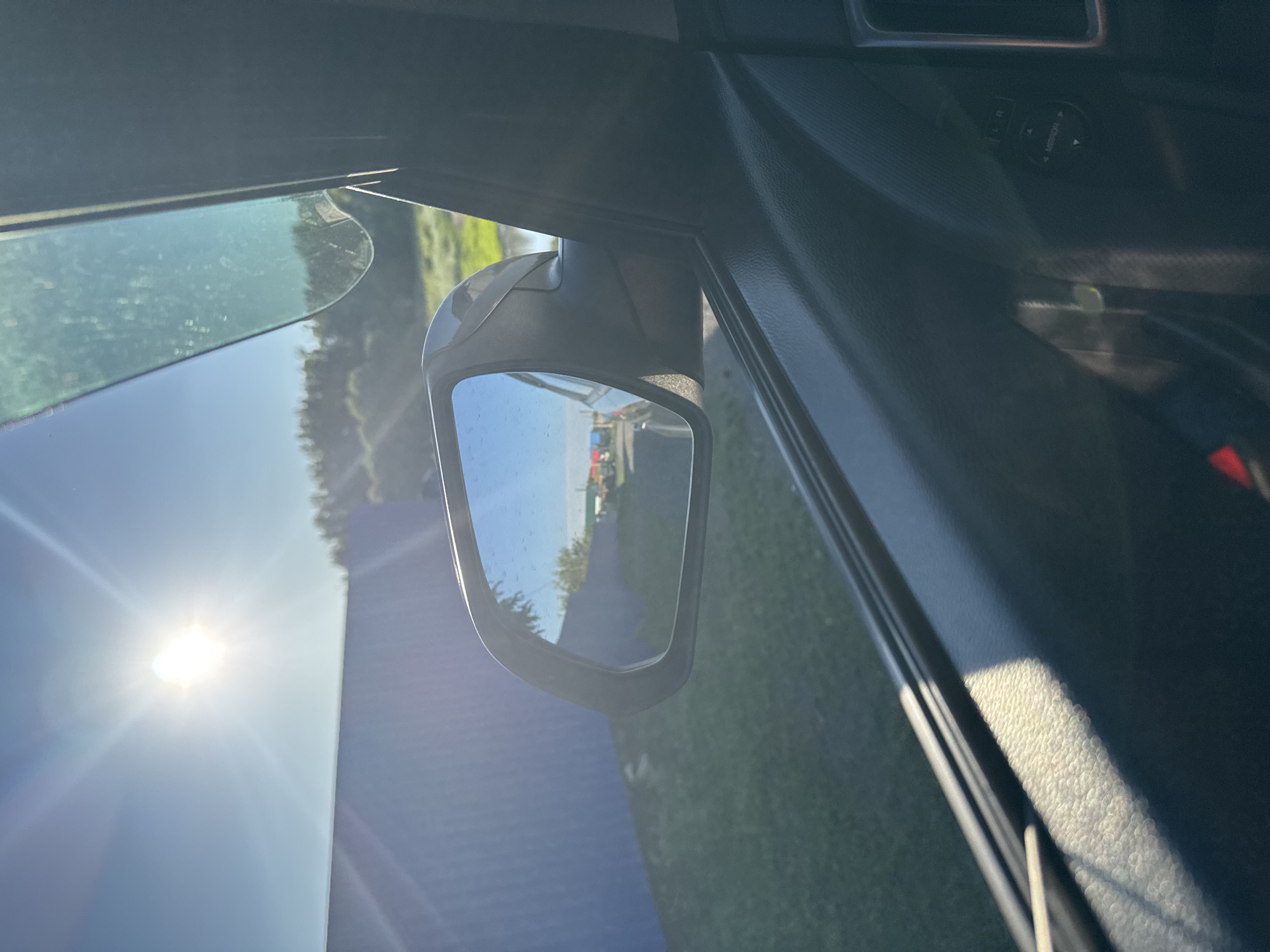 The image is taken from inside a vehicle, looking out the passenger side window and at the side mirror. The sun is shining brightly in the upper left corner of the frame, casting a lens flare. In the distance, visible through the side mirror, is a road with several colorful objects, possibly vehicles or containers, lined up. The side mirror reflects a clear blue sky and some trees. Outside the car, a blue wall or fence runs along the left side of the frame, with green grass in front of it. The interior of the car is visible in the foreground, with the black door panel and a hint of the window controls. The image appears to be taken in a rural or semi-urban environment on a sunny day. The specific location in Starokhalilovo, Russia, is not evident from the visual information provided in the image.