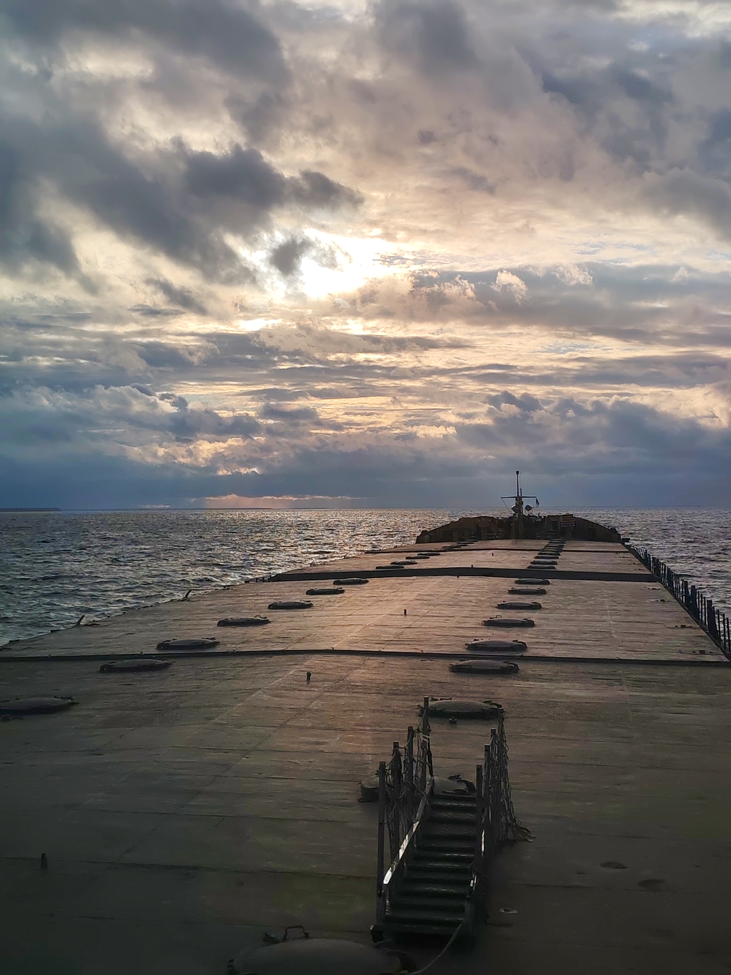 A long concrete pier extends into a choppy body of water under a cloudy sky. The sun is trying to break through the clouds. There are stairs leading up to the pier. The pier has several hatches along its length. In the distance, there appears to be a structure at the end of the pier with some type of antenna on it. The location is Unknown, Unknown.