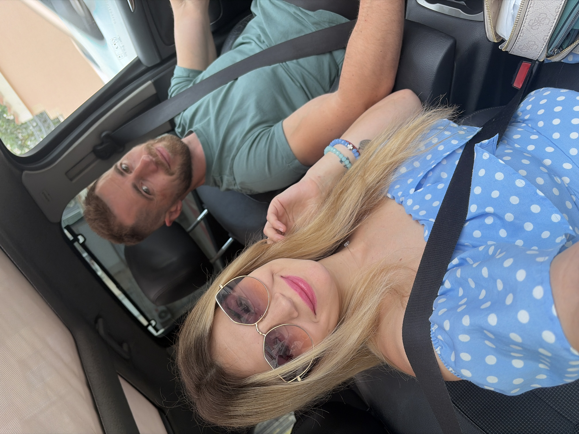 A man and woman are in a car, both wearing seatbelts. The man is in the driver's seat, and the woman is in the passenger seat. The woman is wearing a blue polka dot dress and sunglasses. The man is wearing a green t-shirt. The car has a sunroof. The location is Budva, Montenegro, based on the man's reflection in the sunroof, which shows buildings and vegetation typical of the region.