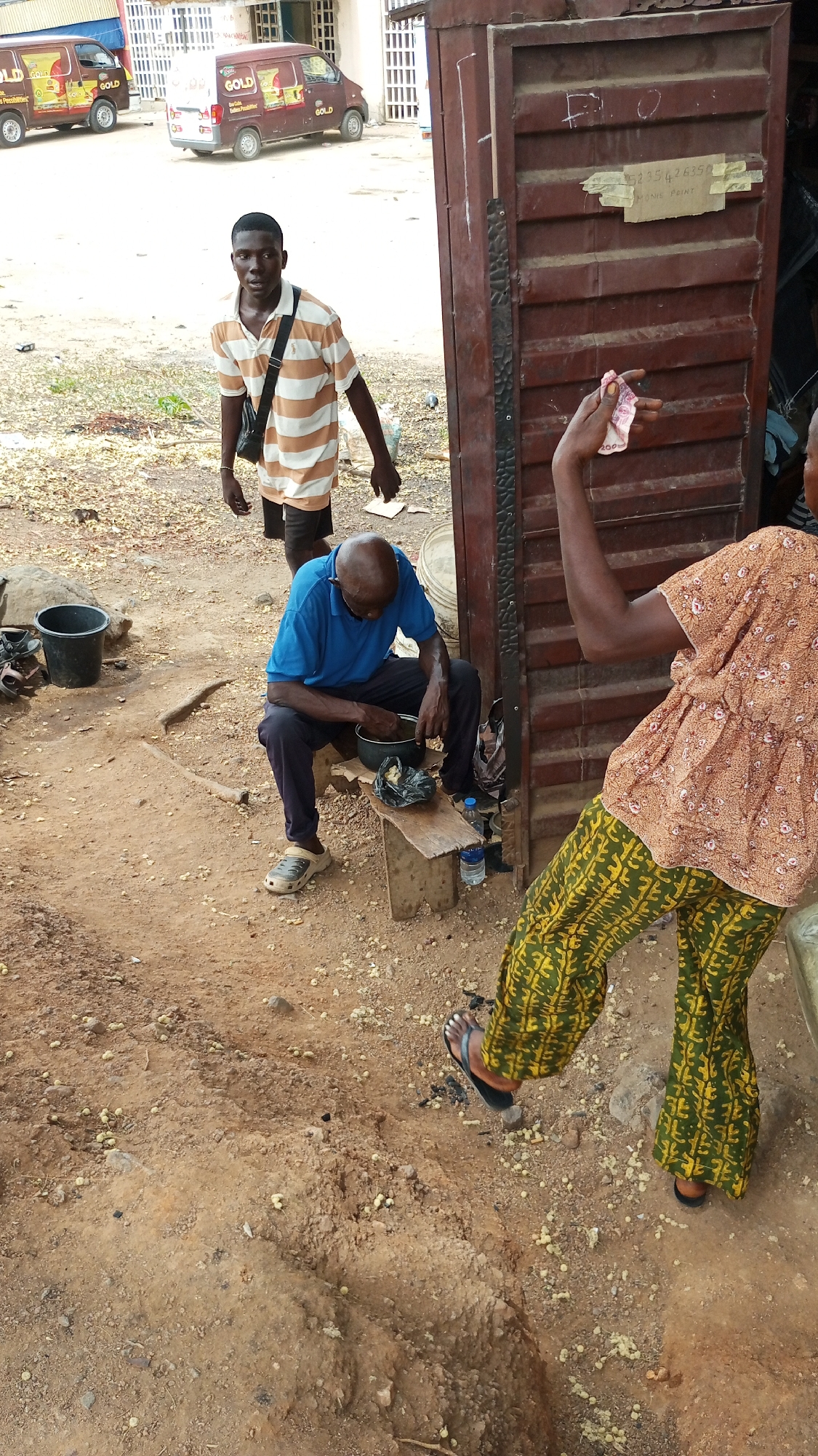 This image captures an outdoor street scene in Lokoja, Nigeria, under bright daylight. The ground is predominantly dirt with scattered small, light-colored pebbles and dry leaves.
Three people are visible in the foreground and midground. On the left, a young man stands, looking directly at the camera. He wears a white and tan striped polo shirt and black shorts, with a black shoulder bag hanging from his right shoulder. Behind him are two brown vans, both branded with "GOLD" and some smaller text, parked on a paved area, suggesting a commercial setting.
In the center, an older, bald man is seated on a small wooden bench. He wears a blue polo shirt and dark trousers, with light grey crocs on his feet. He is bent over, actively working with his hands inside a dark bowl placed on a lower wooden plank. Next to him on the plank is a small black plastic bag and a clear plastic bottle, possibly containing water.
To the right, partially visible, is another person, seen from the back. They wear a light brown patterned top with a ruffled hem and vibrant green and yellow patterned flared trousers. Their right arm is raised, holding what appears to be a crumpled Nigerian Naira note, with "200" clearly visible on it, indicating a 200 Naira denomination. Their feet are in dark sandals.
Behind the sitting man and the partially visible person is a tall, dark brown metallic door or gate, possibly leading into a building or shop. Taped onto this door are two pieces of paper. On the upper one, a phone number "5235426350" is written, and below it, the words "MONIE POINT" are visible, suggesting a financial service point. Black buckets are also visible on the ground to the left of the sitting man. The overall atmosphere suggests a casual, busy market or informal trading area.