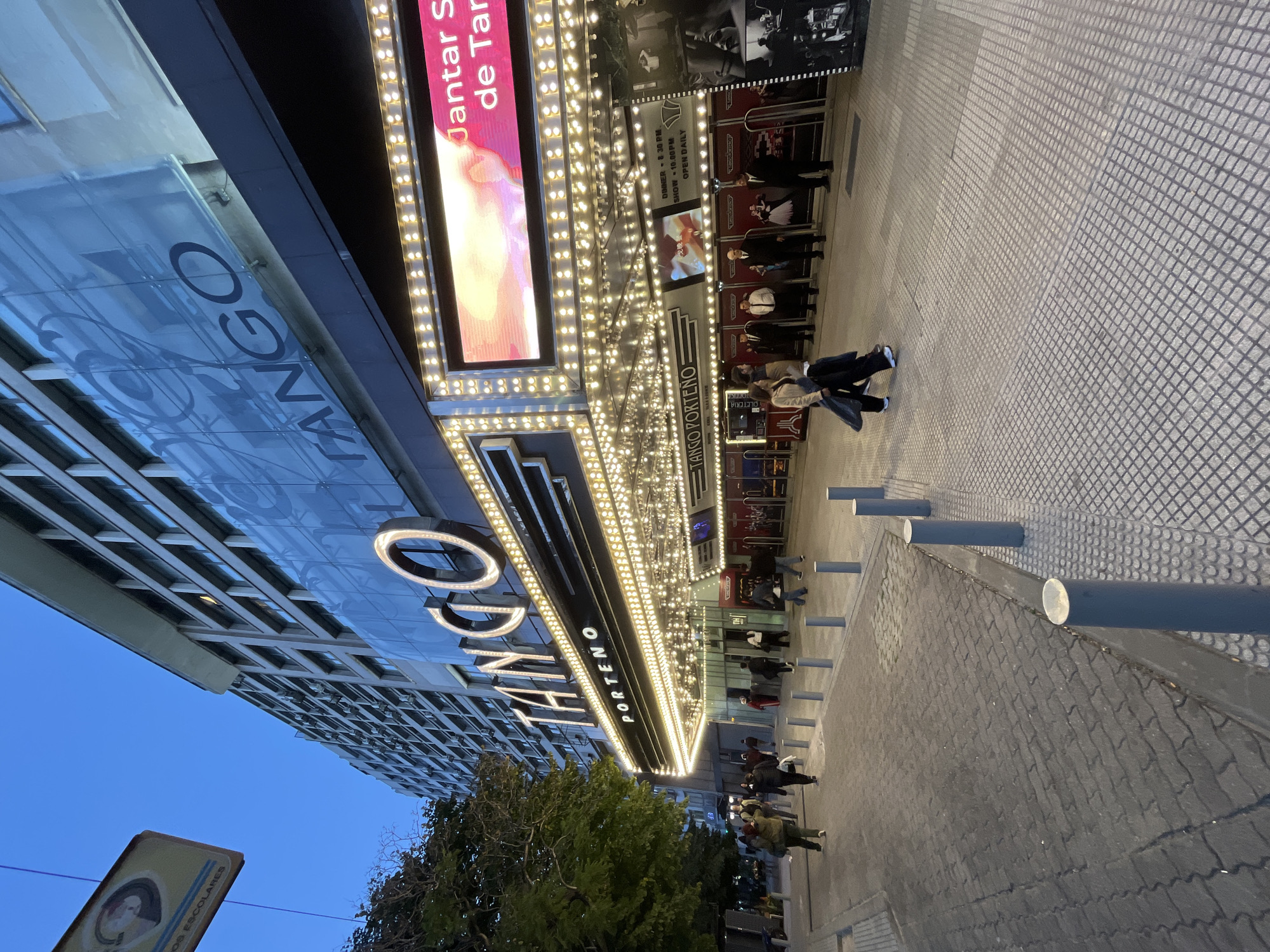 The image shows the exterior of Tango Porteño, a tango show venue in Buenos Aires, Argentina. The building has a prominent marquee with illuminated lettering and numerous light bulbs. A large sign displays "Jantar S de Tar" (likely referencing a dinner and tango show). People are walking on the sidewalk, some near the entrance. Bollards line the edge of the sidewalk. A tree is in the foreground, partially obscuring the lower portion of the building's facade. The sky is visible in the upper left corner. A small sign with an image and the words "OS ESCOLARES" is in the foreground. The paved sidewalk has a grid-like pattern.