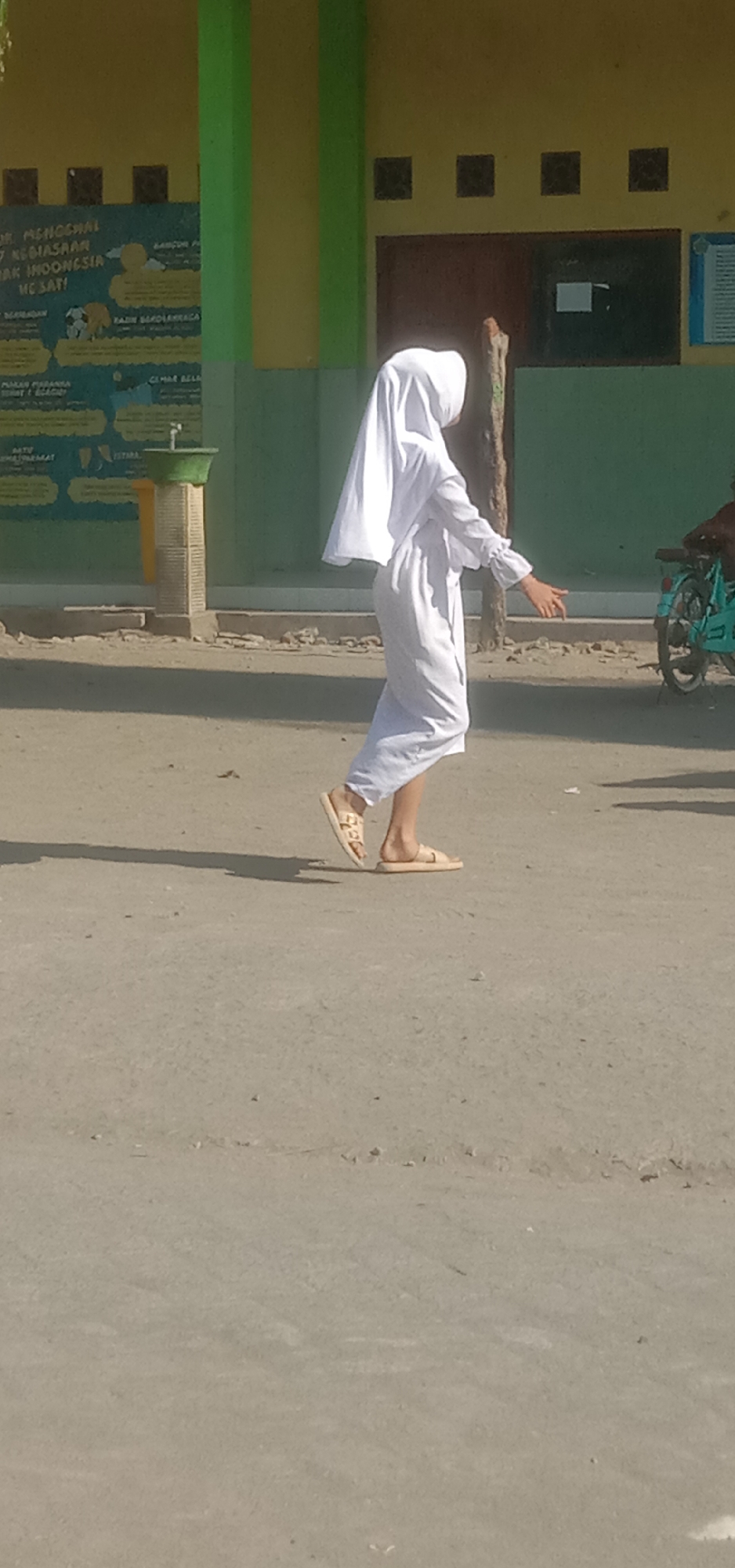 The image captures a young person wearing a white hijab and a long white garment, walking outdoors on a paved surface. The person is positioned in the mid-ground, facing to the left, with their right arm extended forward. They are wearing light-colored sandals. The background features a building with yellow and green accents, and a bulletin board. A tree trunk is visible to the right of the person. In the far right of the image, a portion of a light blue motorcycle is visible. The ground is a dirt or gravel road with scattered debris. The scene appears to be set in a public area, possibly a school campus, given the presence of the building and bulletin board, which is common in Sukorejo, Indonesia. The lighting suggests it is daytime.