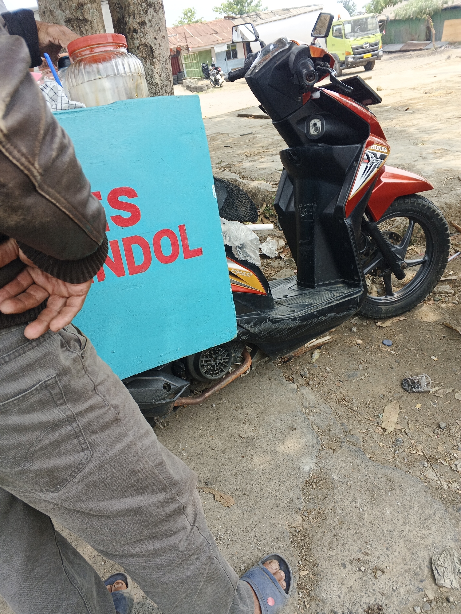 A person wearing gray pants and a brown leather jacket stands next to a bright turquoise box that has red lettering. Part of the lettering reads "MANDOL". A glass jar with a red lid is on top of the box. Behind the box and person is a red and black Honda scooter. The scooter is parked on a dirt and concrete ground. In the background, there is a light green truck and a building with a corrugated metal roof. The scene appears to be in Ubrug, Indonesia.
