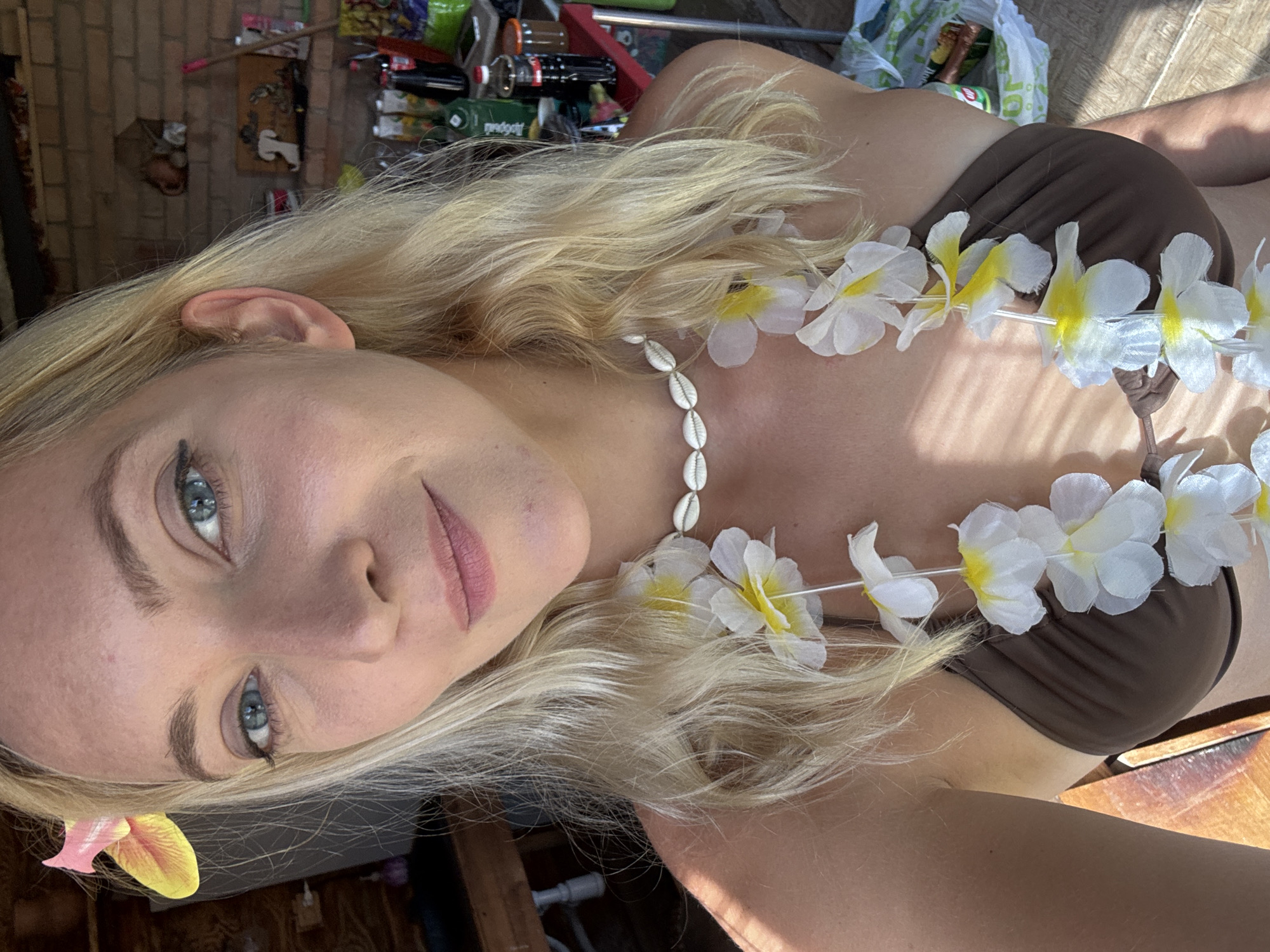 A woman with blonde hair is wearing a brown bikini top and a flower lei around her neck. She has blue eyes and pink lipstick. She is lying down in an outdoor setting. The location may be the city of Дзешчанскі сельскі Савет, Belarus. There is a variety of objects in the background.