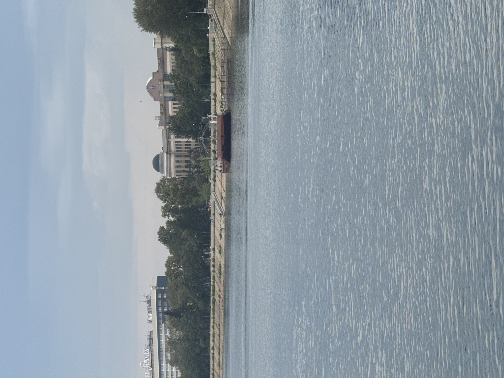 This image depicts a wide, calm body of water, likely a river or lake, with a textured surface reflecting the overcast sky. In the background, a city skyline is visible, characterized by a line of trees separating the water from the urban structures.
Prominently, there is a low, elongated red barge moored near the shore. Attached to the barge and extending towards the land is a dark metal ramp or gangway, and a dark railing system lines the immediate waterfront. Behind the barge, a concrete embankment slopes down towards the water, with visible steps or a ramp leading up to a promenade.
Further inland, a series of buildings form the city's backdrop. To the left, a multi-story white building with many windows displays Cyrillic lettering that reads "ОТЕЛЬ ИРКУТСК". To its right, a larger, tan-colored building with a prominent domed section and a central facade featuring windows and architectural details is visible. This building also has Cyrillic lettering, with "ПРИРОДА" and "КОНЕДИ" legible. Trees of various heights and densities are planted along the embankment and between the buildings.
The sky is overcast with scattered clouds, contributing to a diffused light. The overall impression is a typical urban waterfront scene under a cloudy sky. The presence of the "ОТЕЛЬ ИРКУТСК" hotel clearly places the location within the city of Irkutsk, Russia. There are no clearly identifiable individuals in the image.