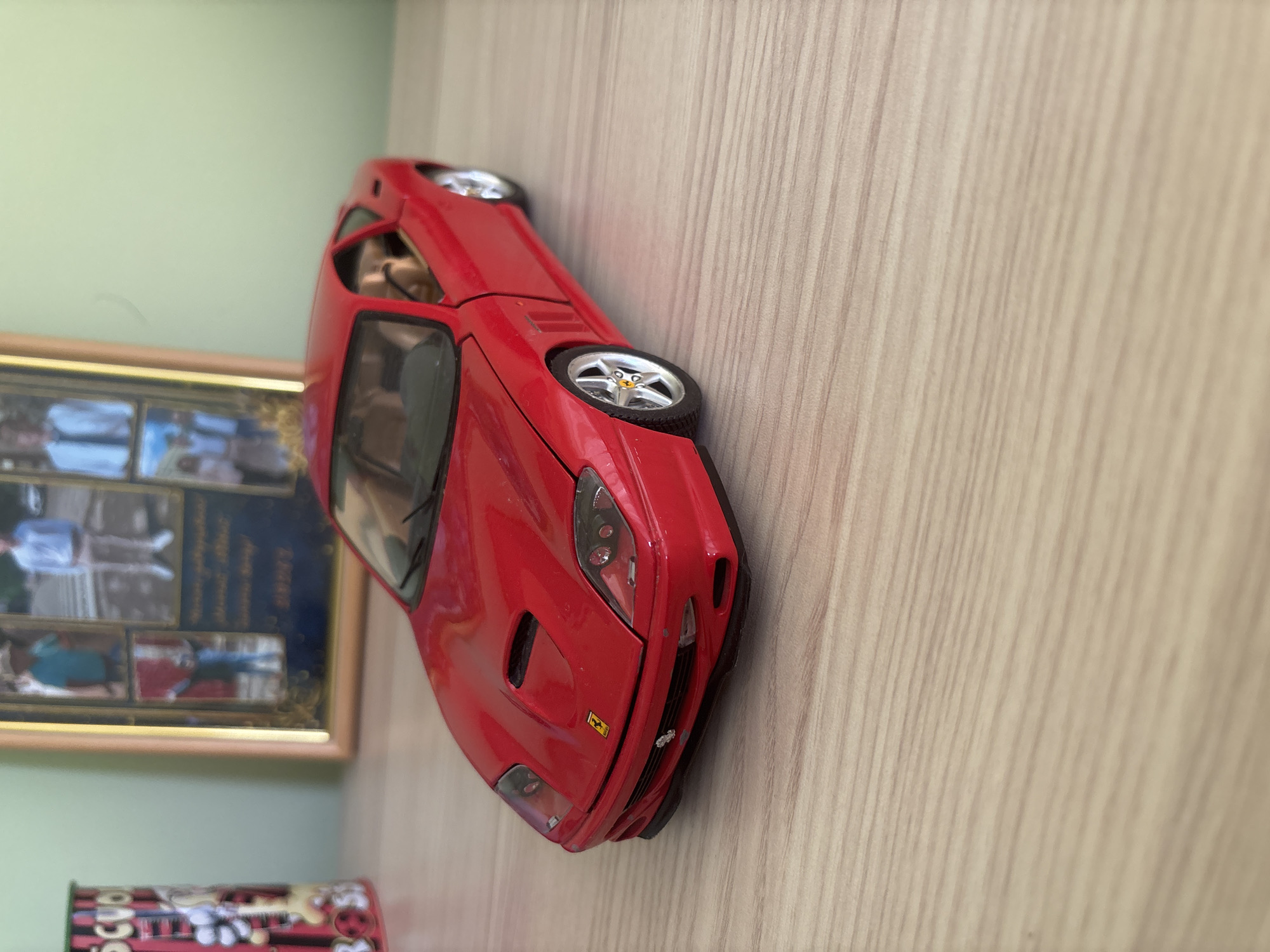 A side-oriented, medium close-up photograph captures a still life arrangement of objects on a light-colored, wood-grained surface, likely a shelf or table, against a pale green wall in an indoor setting.
The primary subject is a vibrant red toy car, specifically a model of a Ferrari, positioned diagonally across the frame. It displays intricate details such as clear headlights, a front grille featuring the yellow Ferrari prancing horse emblem, detailed silver wheels with yellow Ferrari logos on the hubs, and visible tan interior seating. The car's doors appear slightly open, revealing the interior. The wood grain of the surface runs diagonally from top-left to bottom-right, accentuating the car's dynamic placement.
In the background, to the left of the car, a framed picture is partially visible. The frame has a golden or light brown border and encloses multiple smaller photographs, which appear to depict various people. Below the frame, a segment of a cylindrical container or can is seen. Its design includes red, black, and white colors with cartoon-like graphics, possibly a guitar, and faint, illegible text that seems to include "SCVO."
No activity is taking place, presenting a static display of collectible items. The image provides no specific visual cues to confirm the location as Blagoevgrad, Bulgaria. The time of day and weather conditions are not discernible.