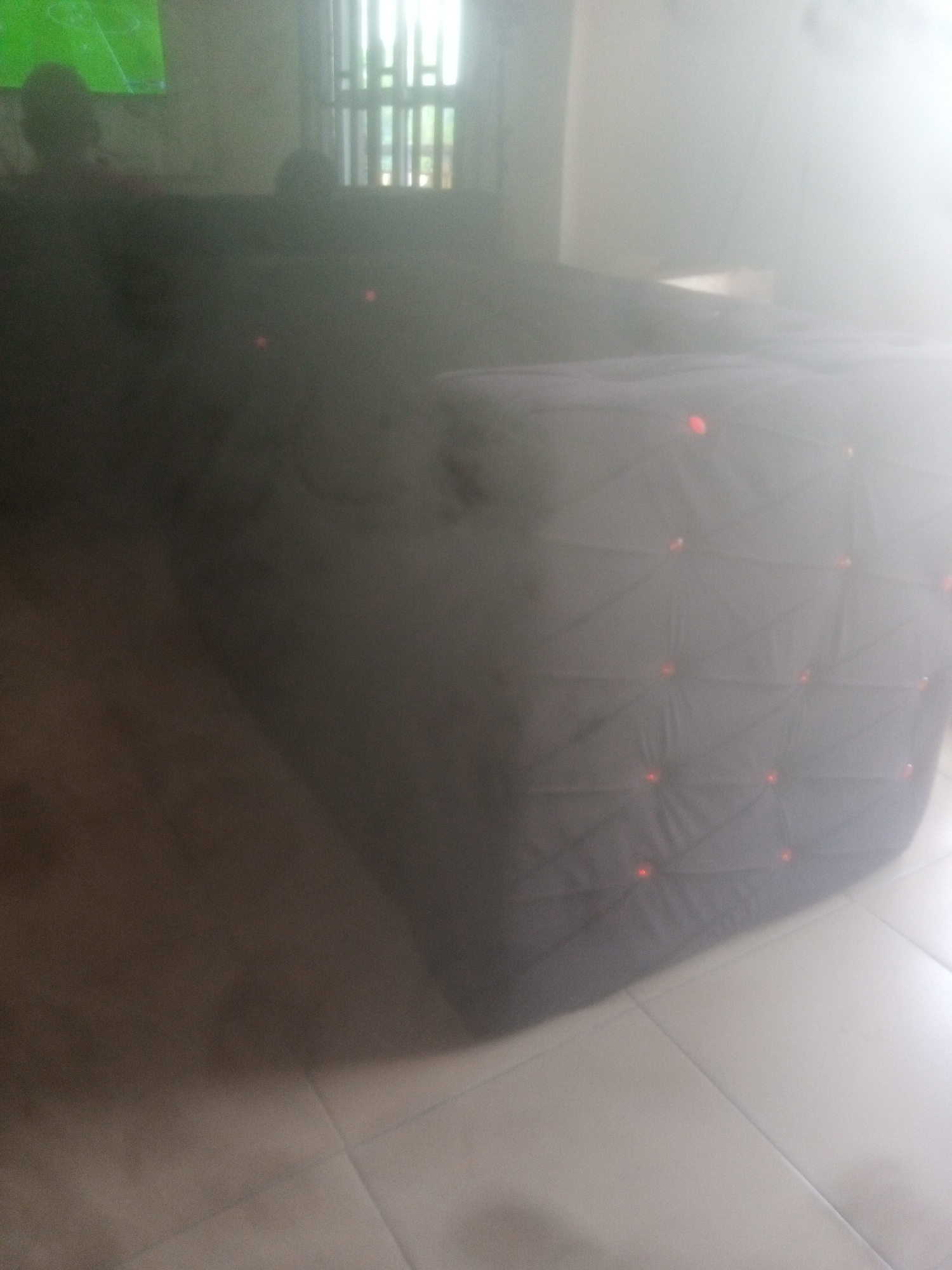 The image depicts a dimly lit room, possibly a living area or a lounge, in Jalingo, Nigeria. The scene is dominated by a large, dark, tufted ottoman or sectional sofa with a geometric pattern and small red lights embedded in it. The lighting is diffused, with a bright light source originating from the right side of the frame, casting a strong glare and creating a hazy effect.
In the background, a television screen displaying a green image, possibly a game or sports broadcast, is visible on the left. Two indistinct human figures, appearing as shadows or silhouettes, are seated in front of the television, suggesting they are watching it. To the right of the television, a window with vertical blinds is apparent, allowing some light to filter in. The floor appears to be tiled. The overall impression is of a casual, indoor gathering.