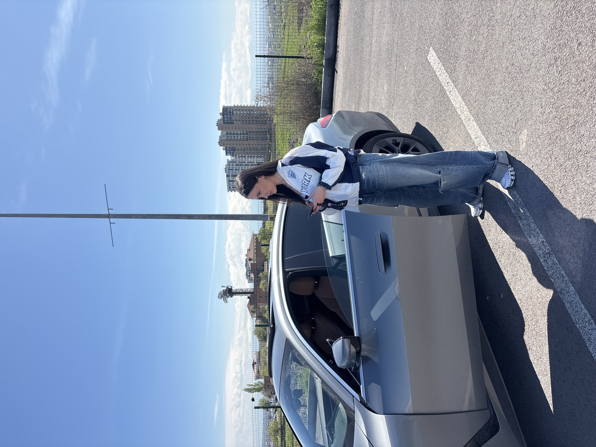 A woman stands next to a silver car in a parking lot, glancing down at a mobile phone in her hands. She is wearing jeans and a white and navy "YORK" jacket. The car door is open. In the background are buildings and a grassy area separated by a fence. A light pole is visible in the foreground. The location is Yerevan, Armenia.