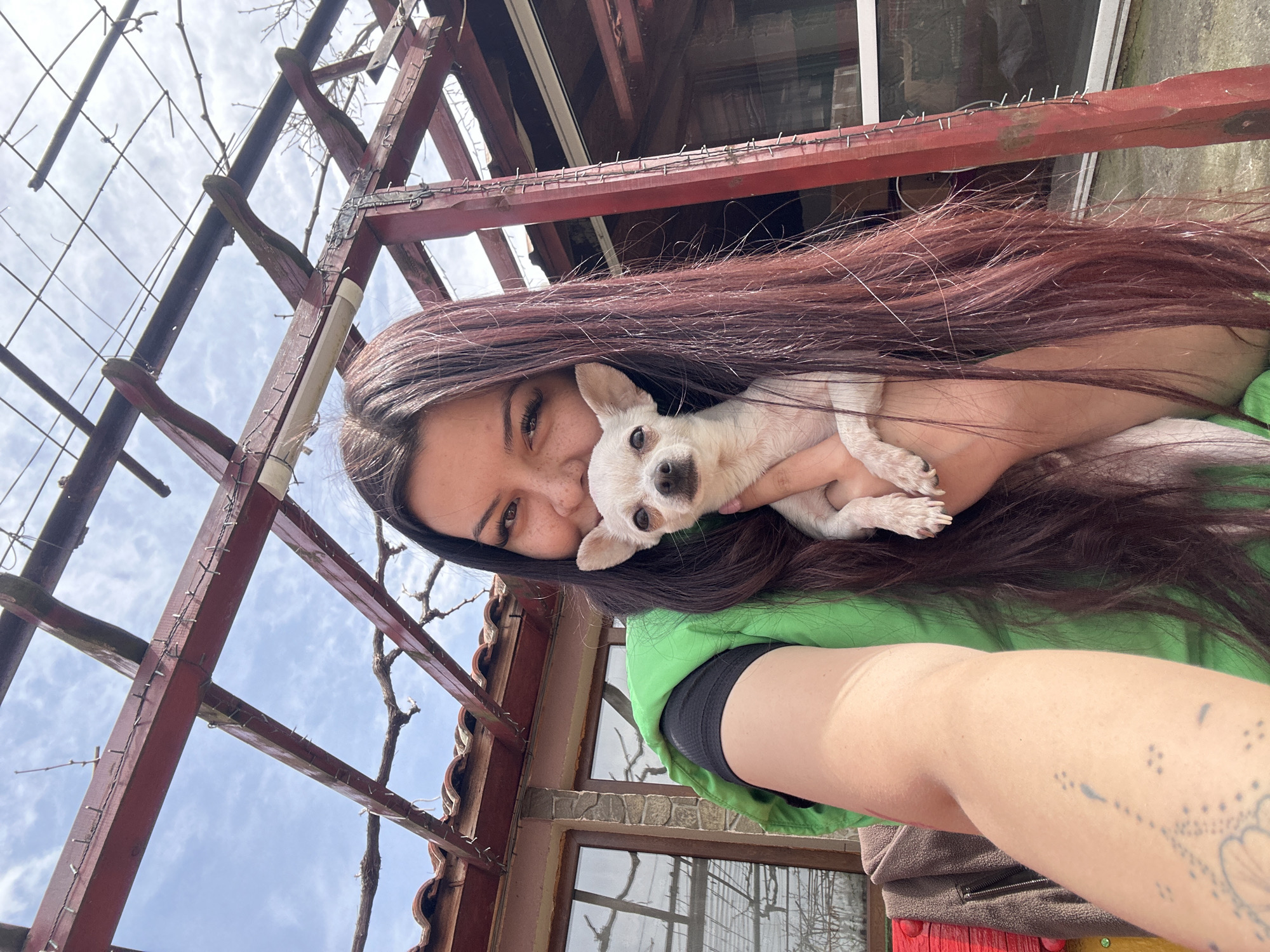 A selfie taken in the city of Chubra, Bulgaria shows a woman holding a small, white dog. The woman has long, dark hair and freckles. She is wearing a green shirt. The dog is looking at the camera. The background includes a wooden pergola and a building. The sky is visible through the pergola.