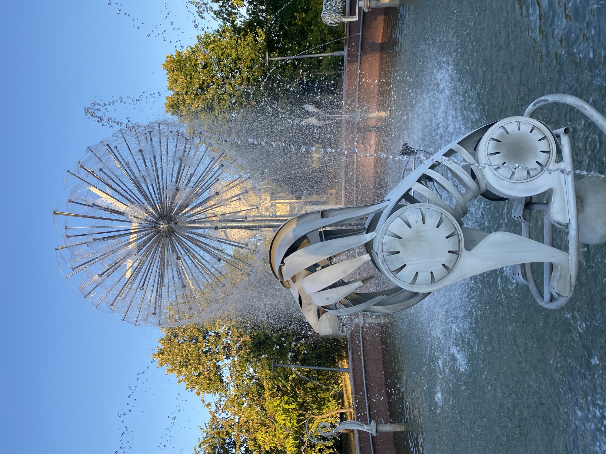 The image depicts a close-up of a contemporary fountain sculpture in Chisinau, Moldova. The sculpture, made of light-colored metal, resembles a stylized animal, possibly a lion, in a crouching or leaping pose. The animal's head is formed from layered, curved metal plates, and its body is a skeletal structure with circular elements that have radiating spokes. The fountain is active, with water spraying from a large, spherical, dandelion-like structure in the background, and smaller jets creating a mist around the central sculpture. The water collects in a pool with a dark, reddish-brown border. Behind the fountain, lush green trees are visible, and the clear sky above is a vibrant blue, suggesting it is a sunny day. The overall scene is one of modern public art and water features, likely located in a park or a prominent public space within Chisinau.