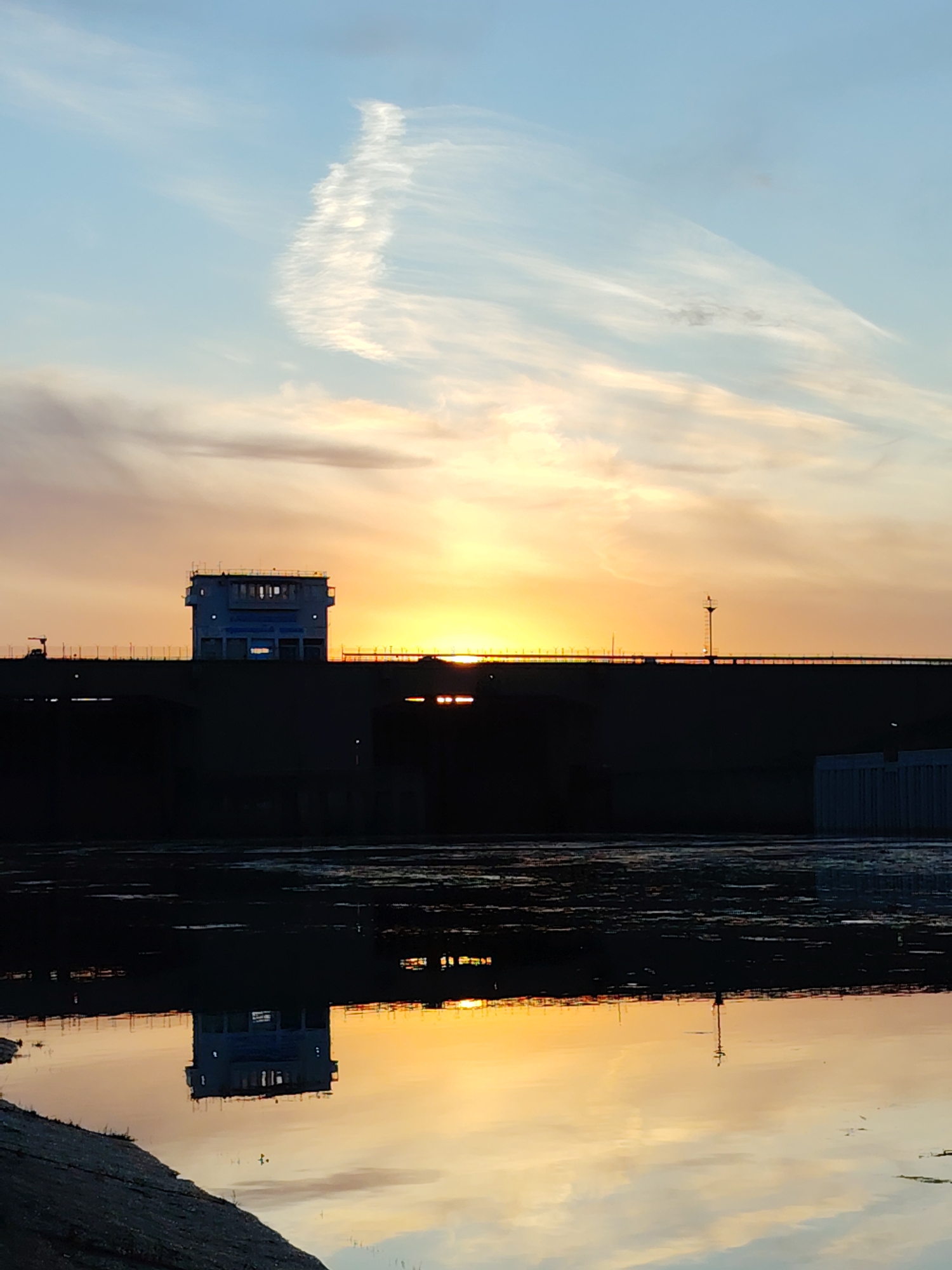 A wide-angle shot captures the Kama River hydroelectric station in Chaikovsky, Russia, at sunset. The water in the foreground reflects the setting sun and the silhouette of the facility. A control building is prominently featured, positioned on a bridge-like structure that stretches across the water. The sky above showcases a gradient of colors from blue to orange, with a contrail streaking across the upper section. The water surface has a layered texture, with dark patches contrasting with the reflections of the sunset. A segment of the riverbank is visible in the lower left corner.