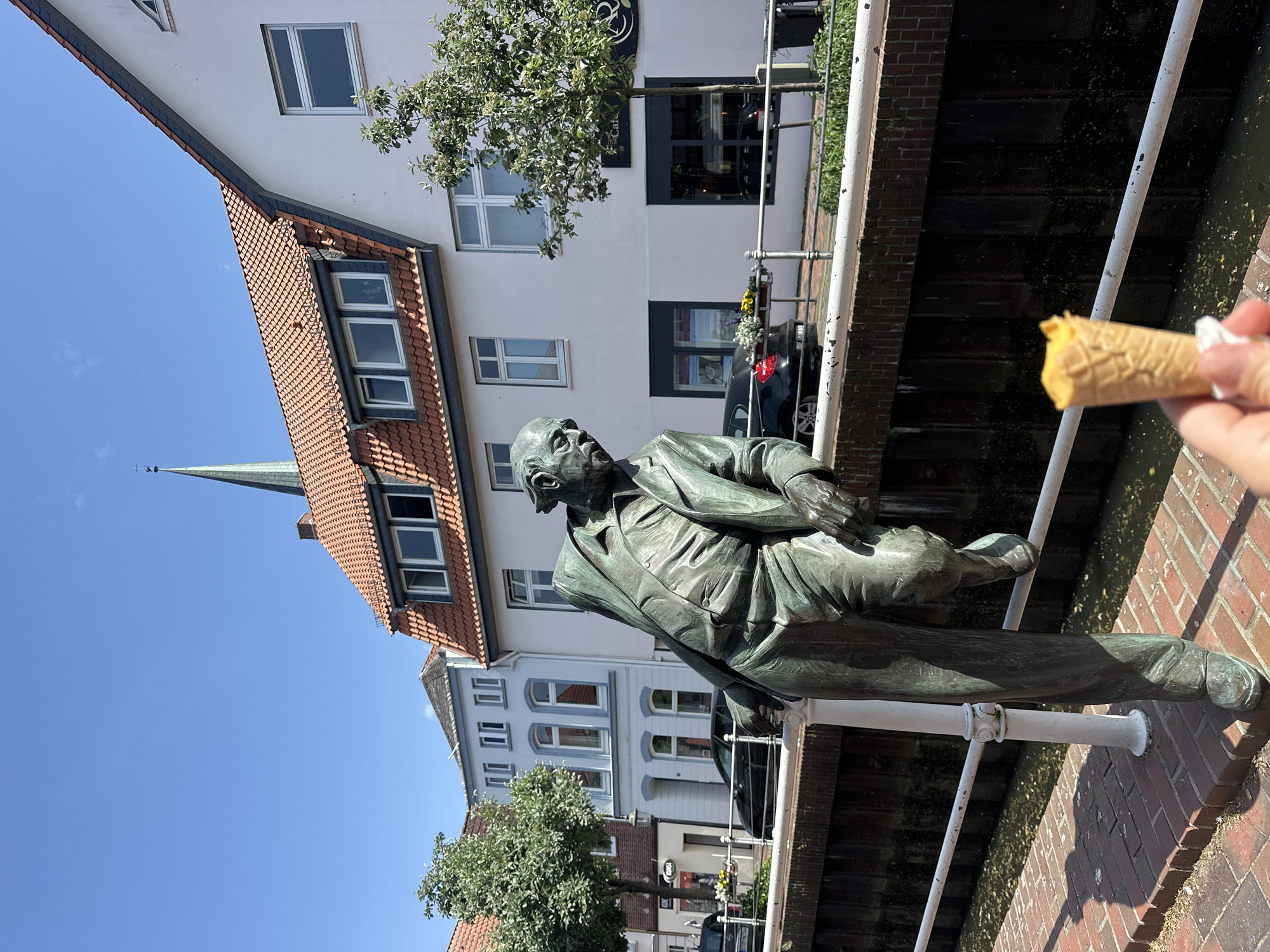 A medium-angle, outdoor daytime shot depicts a bronze statue of a man leaning on a white metal railing, with a building and blue sky in the background. A hand holding a partially eaten ice cream cone is visible in the top-right corner.
The statue is detailed and appears to be of an older man wearing a suit. He is leaning against the railing as if resting or contemplating. The railing itself runs along a brick path or walkway.
In the background, the building is a light color with a red-tiled roof and several windows. A church spire is seen beyond the building. Trees are visible in the middle of the frame and behind the statue. The sky is clear and bright blue.
The location is likely the city of Buxtehude, Germany, based on the architecture and statue style. The statue could be a memorial or artistic installation.