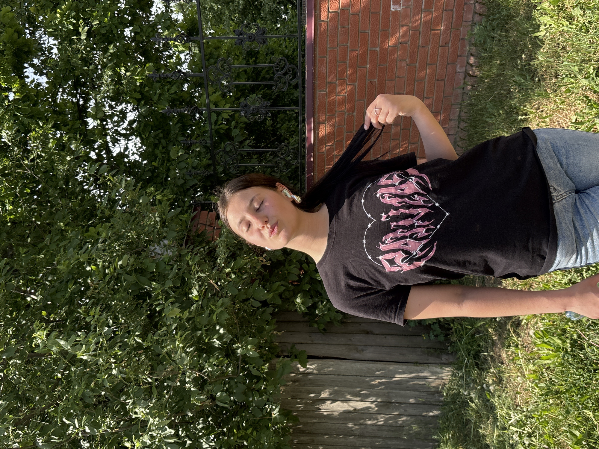 The image depicts a young woman with fair skin, wearing a black t-shirt with a graphic print and blue jeans, standing outside. She has long, dark hair and is wearing what appears to be an earpiece in her left ear. Her eyes are closed, and she is holding a strand of her hair.
The background includes lush green foliage, possibly trees or bushes, and a brick wall. There is also a dark-colored wrought iron fence visible in the background. The ground is covered with grass and what looks like weathered wooden planks.
There is no indication that the location is Фоки, Russia.