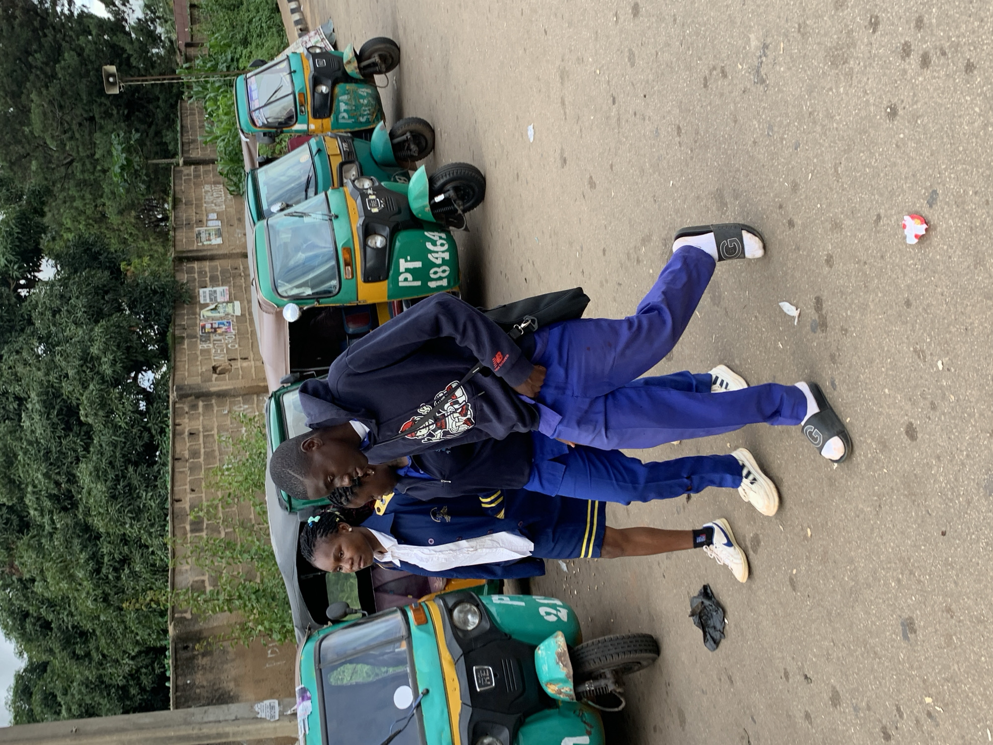 In Barakin Lerawi, Nigeria, three individuals, appearing to be young students in school uniforms, are walking on a paved road. The two in front, a girl and a boy, are looking forward. The girl wears a white collared shirt, a navy blue jacket with yellow trim, and a matching navy blue skirt. The boy wears a navy blue hoodie with a graphic design and purple pants. A third student, a girl, is walking behind them, partially obscured. To their right, a line of three green and yellow auto-rickshaws, also known as "keke napeps," are parked. These vehicles are common forms of public transportation in Nigeria. The background features a brick wall and lush green trees. The ground is asphalt with some scattered debris. The lighting suggests it is daytime.