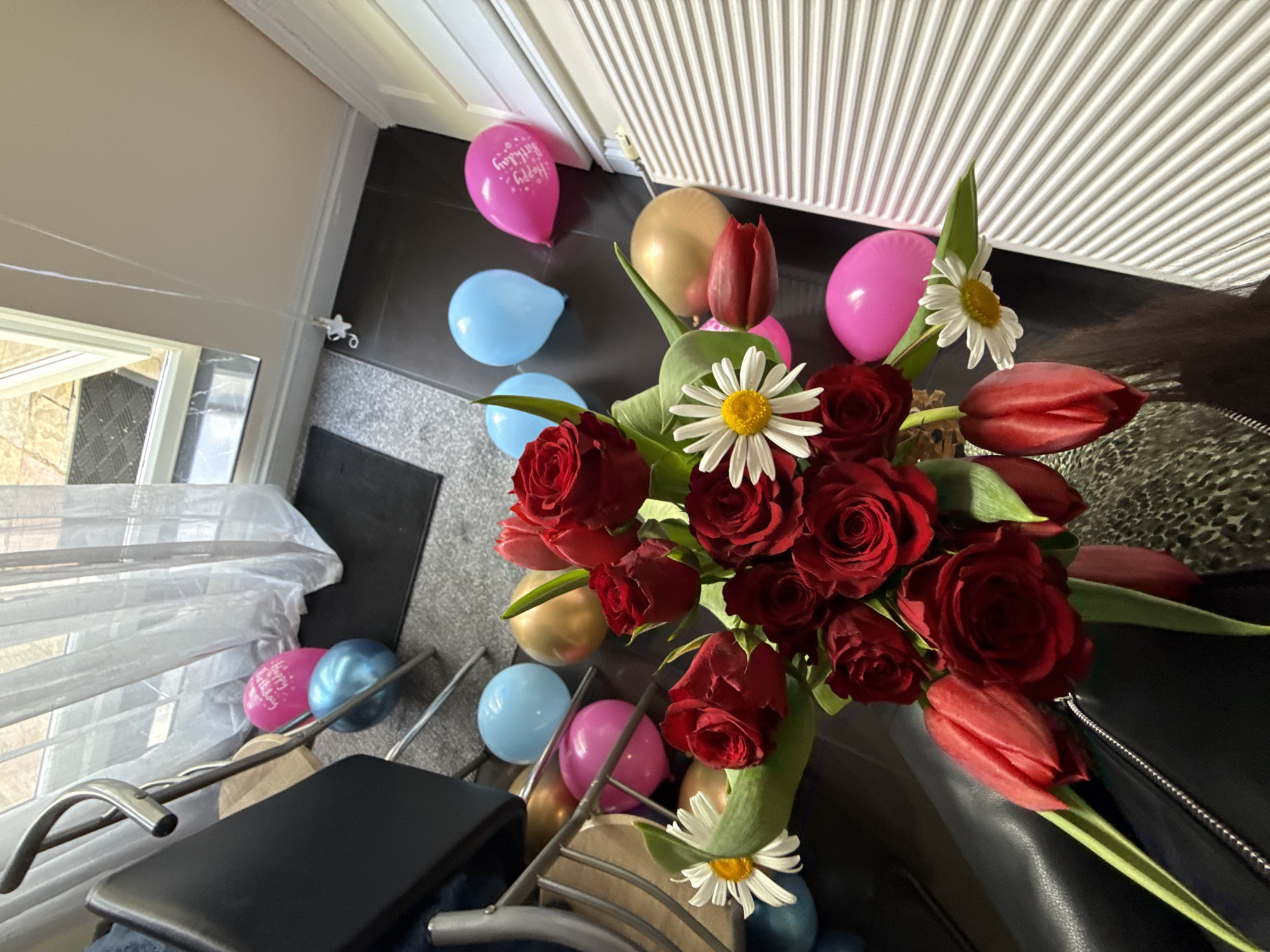 The image shows a bouquet of red roses, tulips, and daisies, with several balloons scattered around. The balloons are in various colors including pink, blue, and gold. One pink balloon has "Happy Birthday" printed on it. The bouquet is positioned near a black chair, and the background shows a window with curtains and a section of a louvered door or wall. A part of the wall is painted light gray. The location appears to be indoors. I have no evidence to confirm the location as Great Yarmouth, United Kingdom.