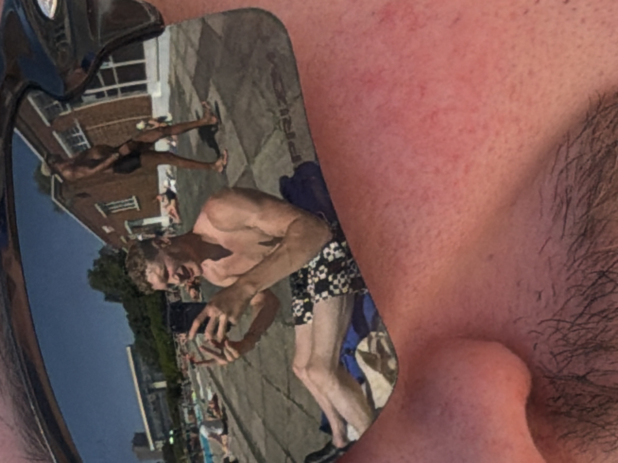 A close-up shot of a pair of sunglasses reflecting a scene in what appears to be London, United Kingdom, next to a person's face. The sunglasses reflect a shirtless individual taking a photo with a smartphone in the foreground, wearing black and white shorts and black shoes. A building and a group of people in swimwear can be seen behind the person.