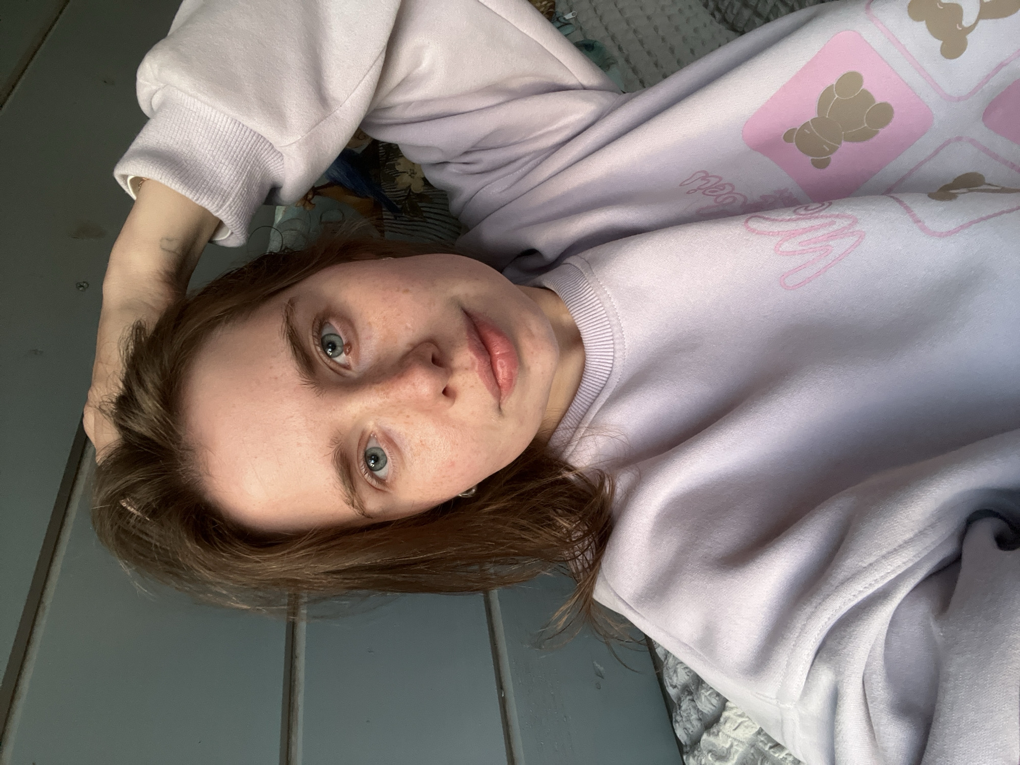 A young woman with light brown hair and blue eyes is lying down, wearing a lavender sweatshirt with a design of pink squares containing teddy bears. Her left arm is raised and her hand is near her head. She is indoors; to her left is a grey, slatted surface, possibly a bed frame or wall. To her right is a textured, gray and white fabric, presumably part of the bedding. The photo appears to be taken from an above perspective. The location is not specified in the image.