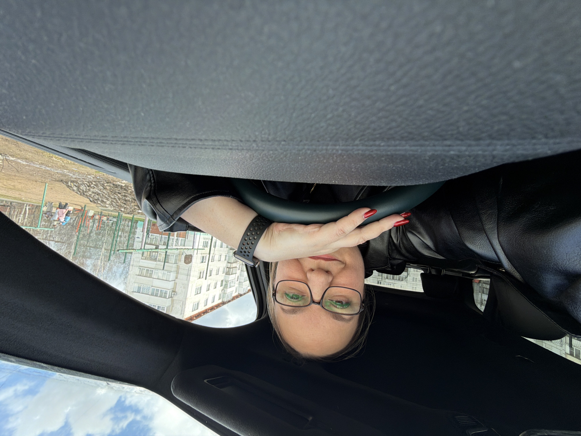 A woman is captured in a selfie taken from inside a car, looking upwards towards the camera. She is seated in the driver's seat, with her head and upper torso visible. She wears glasses, a black leather jacket, and has red nail polish on her fingers. A black smartwatch is visible on her left wrist, as her left hand rests on the steering wheel. The car's interior, including parts of the steering wheel, dashboard, and roof lining, is dark-toned.
Through the car's windshield and side windows, a residential area in Chaikovsky, Russia, is visible in the background. Several multi-story apartment buildings with numerous windows are present. The trees are bare, suggesting late winter or early spring. Patches of brown ground and some green fencing, indicative of a playground, are also discernable. The sky is partly cloudy with patches of blue. The overall setting is a daytime scene, with natural light illuminating the surroundings. The woman maintains a neutral expression, looking directly at the camera. There is no visible text in the image.