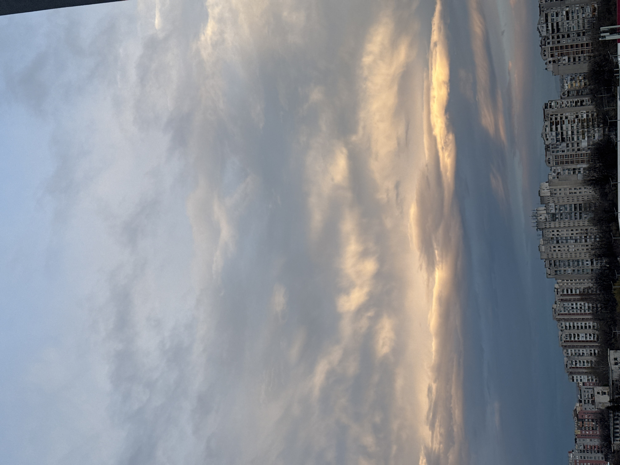 The image depicts a view in Plovdiv, Bulgaria, featuring a row of tall, multi-story apartment buildings along the right side. The sky occupies most of the frame, showcasing a mix of clouds ranging from light gray to dark blue. Some clouds are illuminated with a golden hue, likely from the setting or rising sun. A dark, indeterminate shape is visible in the upper-left corner of the image, possibly part of a building or structure from where the photo was taken. Trees are interspersed among the buildings, but details are obscured. The overall impression is of a cloudy urban landscape at dawn or dusk.