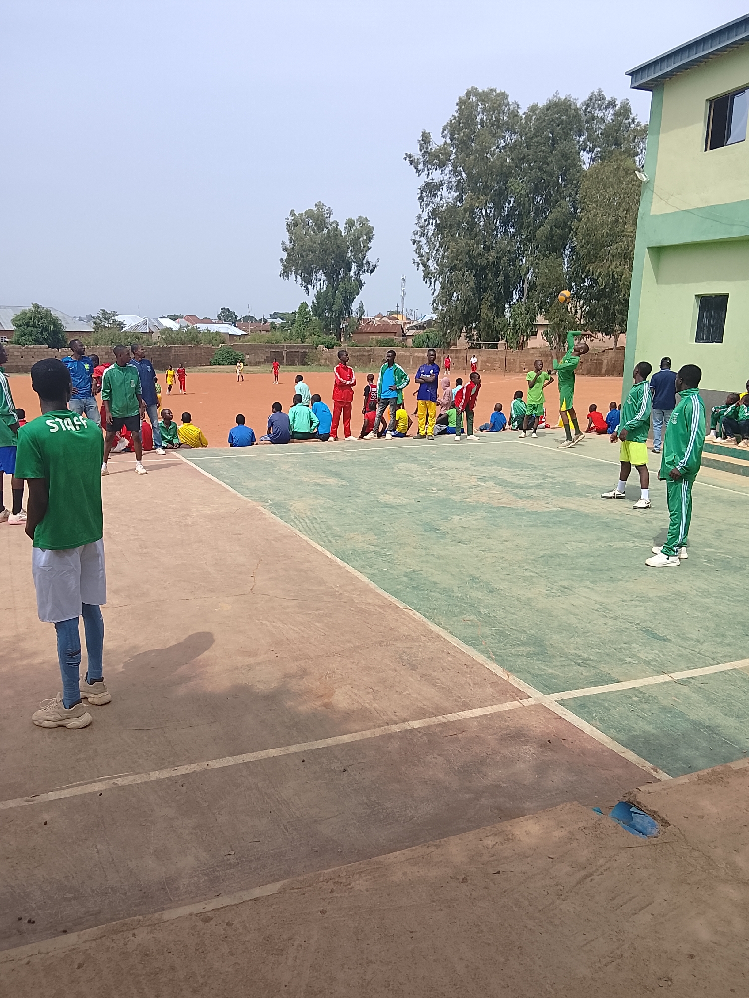 A wide-angle shot depicts an outdoor scene likely in Gura Top, Nigeria, on a hazy day. A group of people, mostly young men and teenagers, is gathered on a worn, multi-toned green and brown cement court marked with white lines.
The court appears to be used for sports, as one young man in a green uniform is captured in the middle of a jumping motion with a yellow ball in his hand, presumably throwing it. Others in similar green uniforms are standing around, seemingly observing or participating in the activity. Several individuals are seated or standing along the perimeter of a red clay area adjacent to the court, some wearing colorful clothing.
In the foreground, a person wearing a green shirt with the word "STAFF" printed on the back stands facing away from the camera. Trees are in the background and a two-story green and beige building is on the right side of the frame. The ground in the foreground is a darker, rough cement surface.