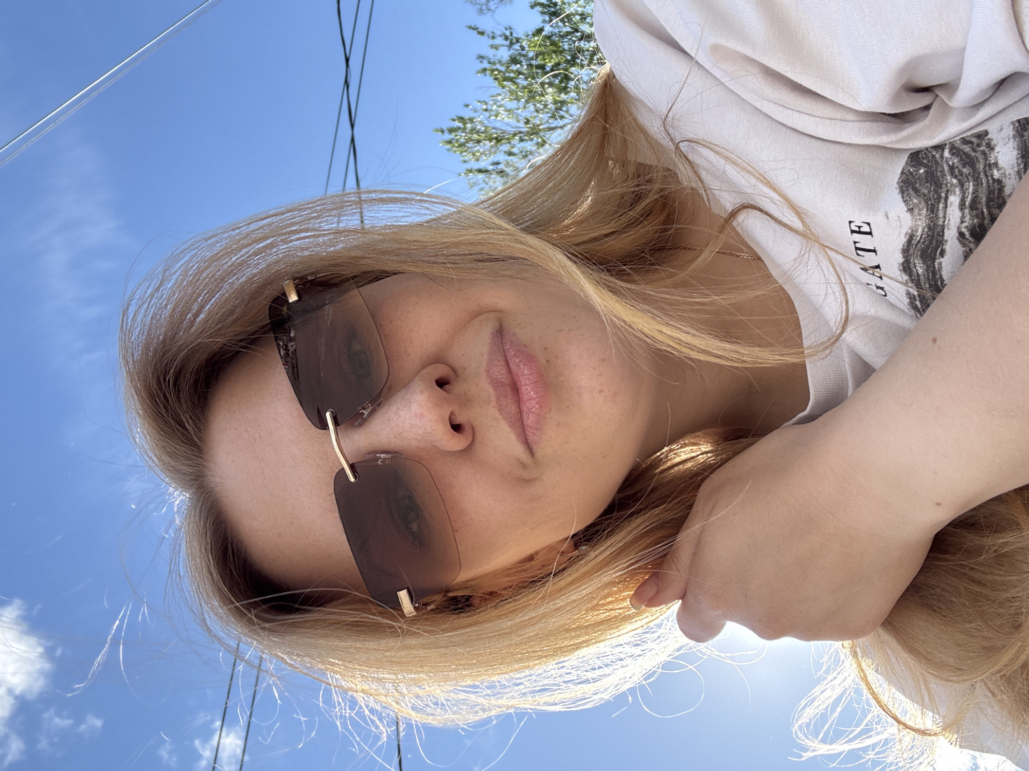 A light-skinned woman with blonde hair, wearing frameless sunglasses and a white hooded sweatshirt with printed text, is looking directly at the camera. The photograph is taken from a low angle, with a clear blue sky visible in the background, along with power lines and foliage. The woman's left hand is visible near her face. The photo's location cannot be verified based on image contents.