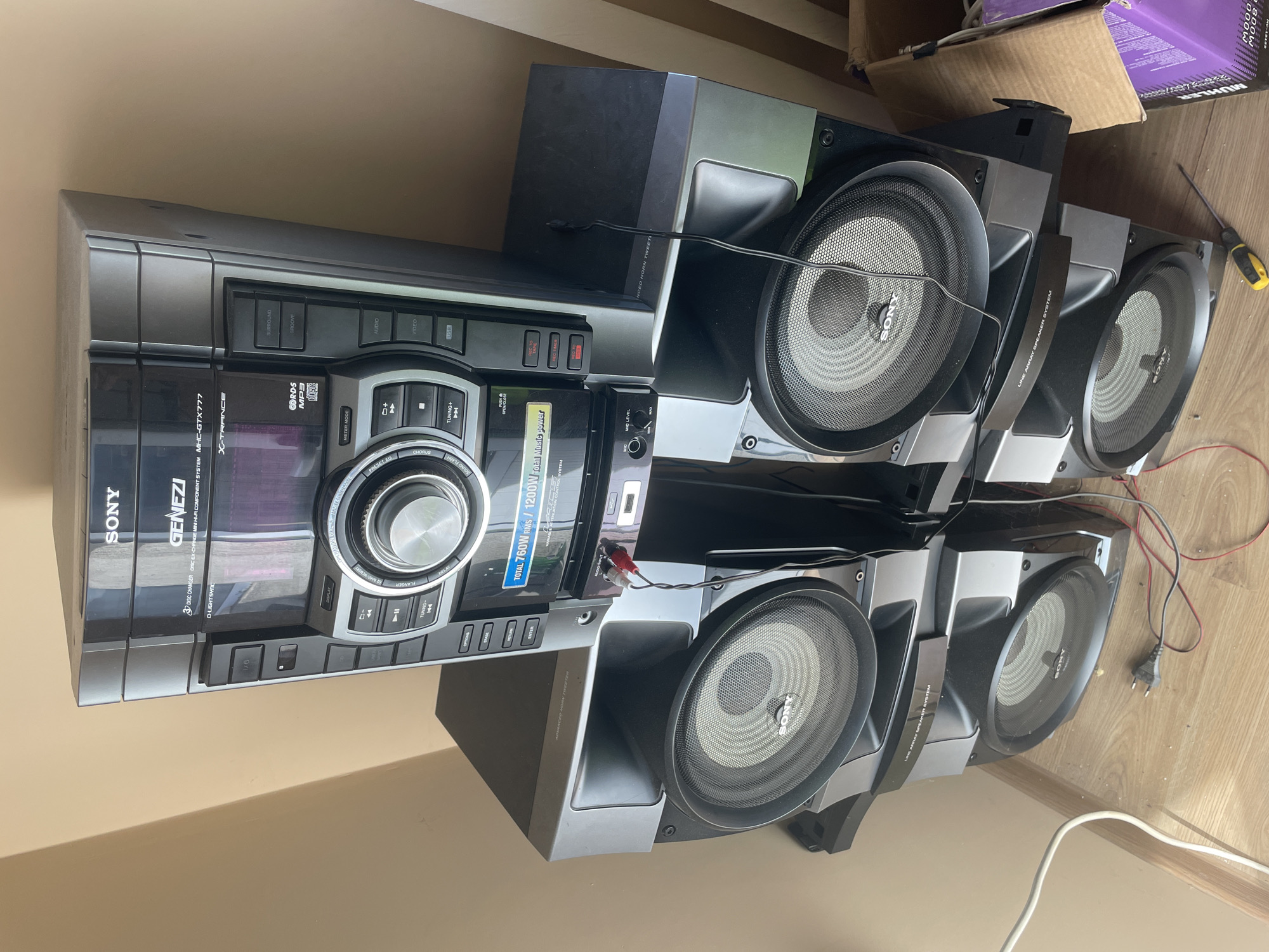 The image displays a Sony Genezi mini hi-fi component system, model MHC-GTX777, positioned indoors against a beige wall on a wooden floor, suggesting a domestic setting in Ihtiman, Bulgaria. The system comprises a main unit and multiple large speakers.
The central unit is black and silver, featuring a "DISC CHANGER" that supports "CD-R/RW MP3 disc," and an "X-TRANCE" audio enhancement. Visible controls include a large central dial, buttons for "SURROUND," "GROOVE," "AUDIO," "VIDEO," and "USB" inputs. It also has "TUNING+/-", "RESET EQ," "CHORUS," "SOUND FLASH," and "OPTIONS" buttons, along with a "METER MODE" and "D-LIGHT SYNC" feature. The power output is prominently displayed as "TOTAL 760W RMS / 1200W Total Music power." Microphone inputs with "MIC LE-VIL" controls are also present.
The speakers, branded "SONY," are part of a "LINE ARRAY SPEAKER SYSTEM" and incorporate "ADVANCED HORN TWEETER" technology. They feature large cones and are connected to the main unit by various black and red audio cables. A white power cable and a European-style two-pin plug are visible on the floor. In the background, a cardboard box with the text "MUHLER," "MOOO," and "8" is visible, along with a yellow-handled screwdriver. No specific activity is depicted; the system appears to be in a state of being set up, moved, or stored.