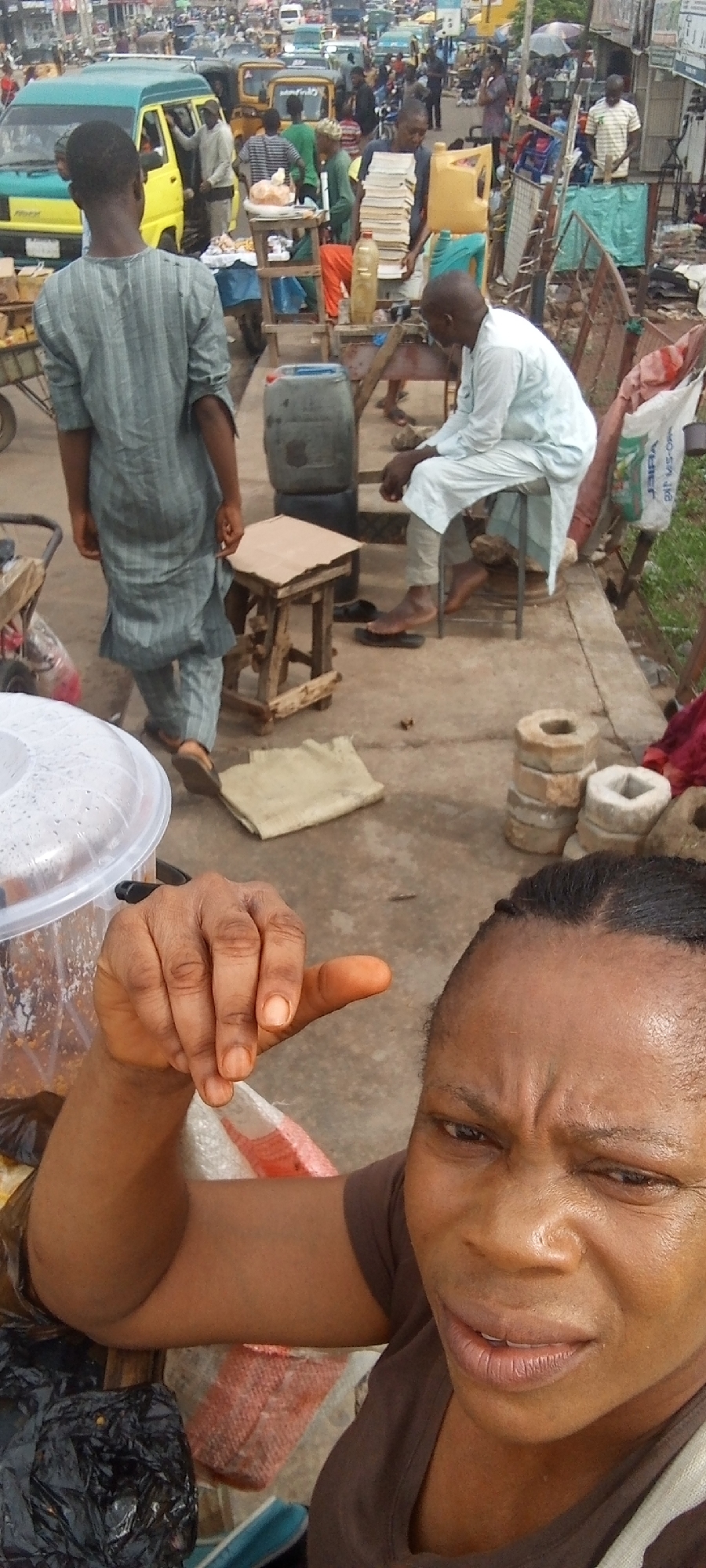 A congested urban street scene in Maiburiji, Nigeria is captured in the photograph. The foreground features a person making a dismissive gesture with their hand. Various people in the background are walking and standing near vehicles, including cars and buses. Merchandise is displayed on tables and carts, suggesting a market setting. A man sits on a stool near a large container. Other people are walking by. The overall environment appears busy and densely populated.