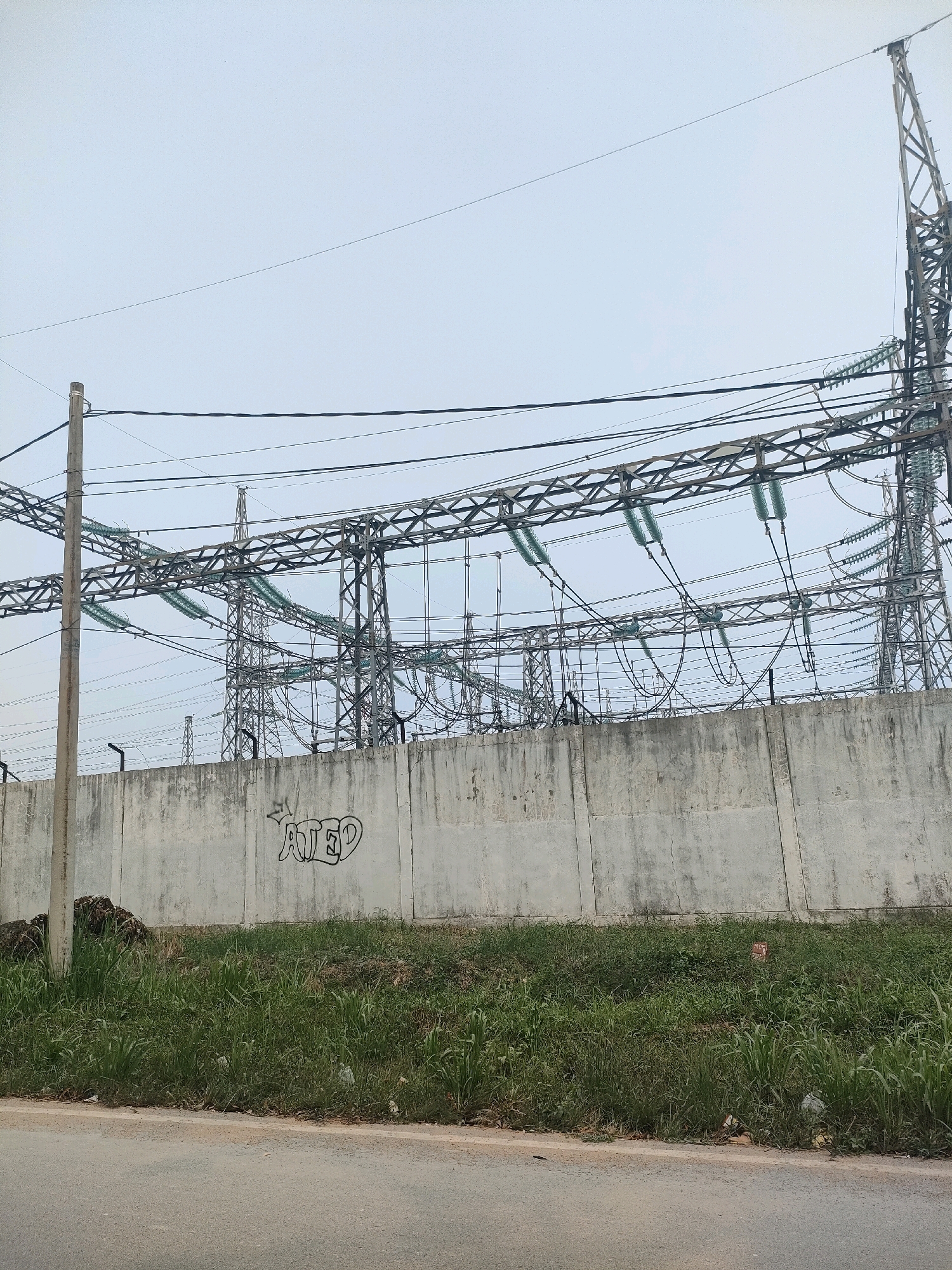 This image depicts an outdoor scene in Batam, Indonesia, focusing on an electrical substation. The foreground shows a paved road, followed by a strip of green grass and vegetation. Behind the grass is a tall, weathered concrete wall, which serves as a barrier for the substation. The wall has some graffiti on it, including the word "ATED" in stylized lettering.
Beyond the wall, the substation itself is prominent. It features a complex network of steel structures, high-voltage power lines, and insulators, all against a pale, overcast sky. Several tall, lattice-like towers support the numerous electrical cables. The overall impression is one of industrial infrastructure, with the natural elements of grass and sky contrasting with the man-made structures. The image appears to be taken during the daytime, likely under cloudy conditions, as there are no strong shadows.