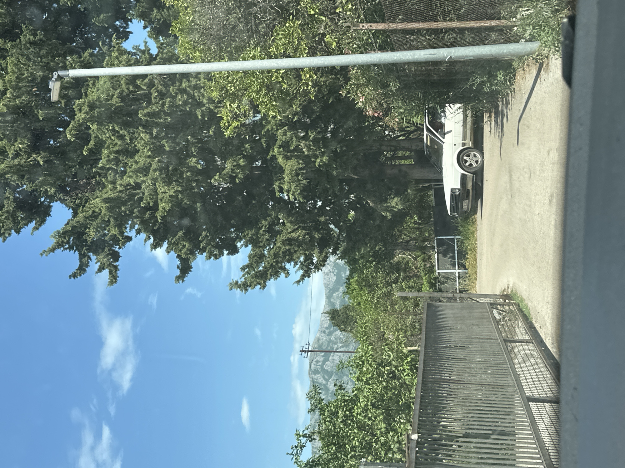 The image, likely taken from inside a vehicle, shows a view in Bar, Montenegro. The sky is blue with scattered clouds, and a mountain range is visible in the distance. A large evergreen tree dominates the left side of the image, obscuring some of the sky. In the middle ground, there is a metal fence running parallel to the road. To the right of the fence, a white car is parked on a paved area. A utility pole is noticeable in the background. The immediate foreground shows a partial view through a window, with elements of the vehicle's interior visible on the right side of the frame. The scene appears to be a residential area.