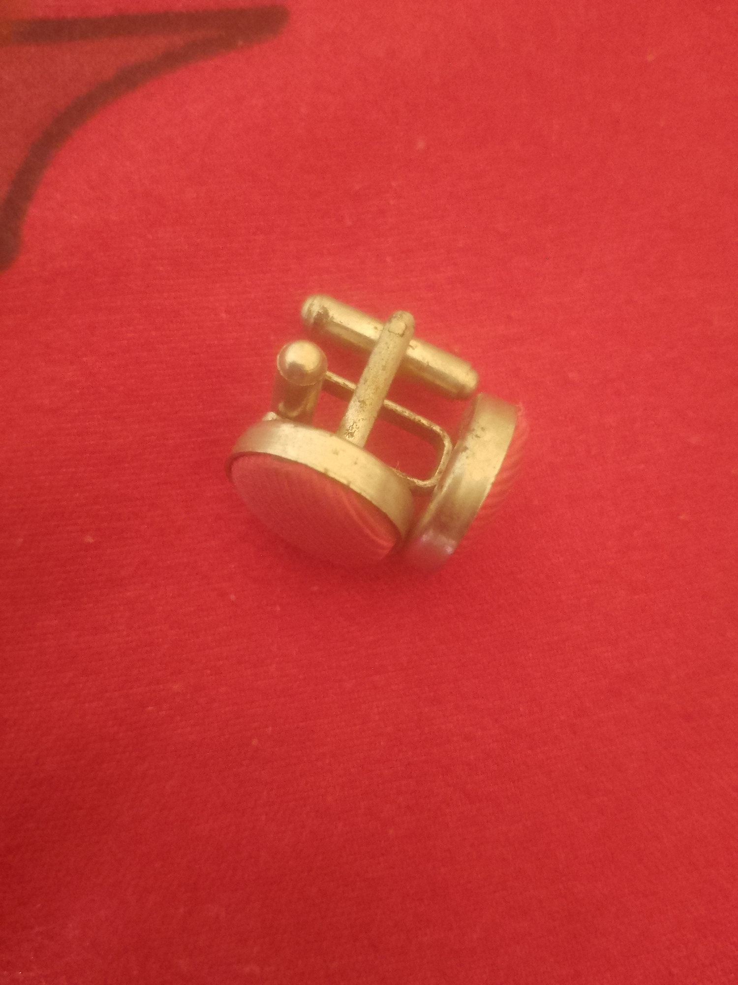 A close-up, high-angle shot shows a pair of gold-toned cufflinks with pinkish-red, oval-shaped inlay stones, resting on a textured red fabric. The cufflinks are angled, with the barrel mechanism of one visible. The fabric appears to be velvet or a similar soft material, with a slight sheen. The background is entirely red fabric, with a shadow in the upper left corner suggesting an unseen object. The scene is static and does not depict any activity or individuals. The location is not discernible from the visual information provided.