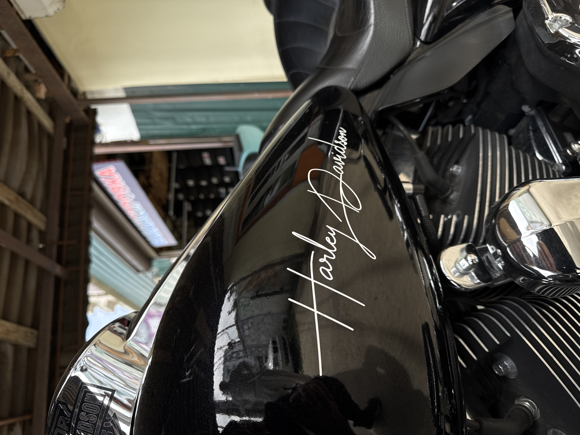 The image shows a close-up of a black Harley-Davidson motorcycle, with the iconic Harley-Davidson logo prominently displayed. The shot is taken in an urban environment, with glimpses of buildings and structures visible in the background. A store front in Krasnodar with a sign is partially visible. The motorcycle is the central subject, with its glossy black finish and chrome accents reflecting the surrounding environment. The angle suggests the photo was taken from a slightly elevated position. The overall impression is one of urban life and motorcycle culture.