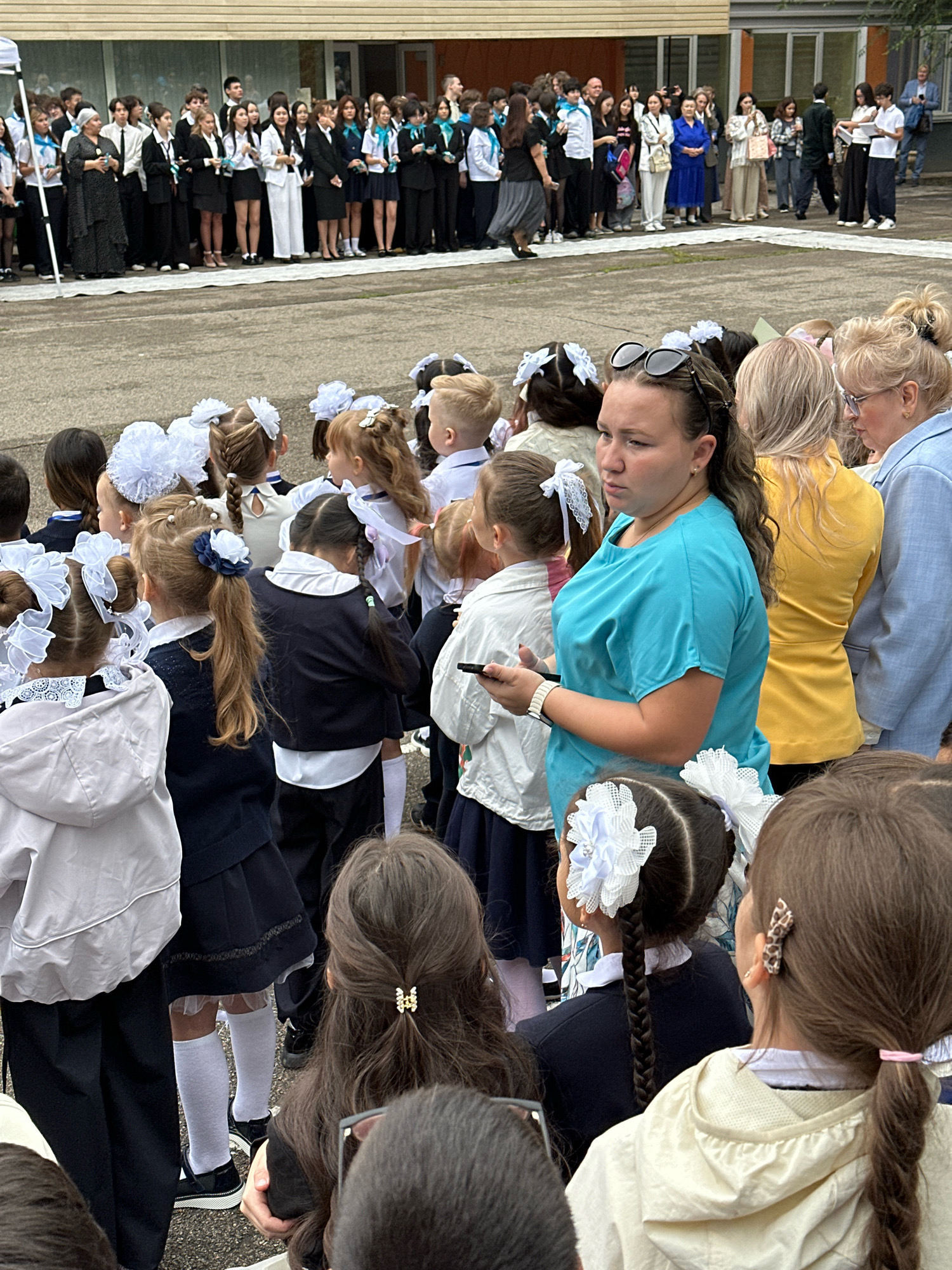 An outdoor event is taking place in front of a building with a light brown facade. The ground appears to be asphalt. A crowd of people are gathered, with many children in school uniforms. The children are wearing dark blue skirts or trousers and white shirts, some with white bows and ribbons in their hair. Adults are also present, some wearing formal attire. The weather appears to be overcast. The scene is likely a school event, possibly the first day of school, given the attire of the children. The architecture suggests a location in Almaty, Kazakhstan.