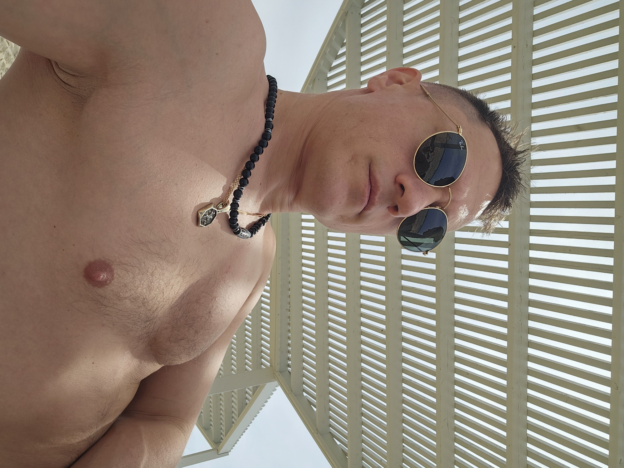 A shirtless man with sunglasses and a necklace stands under a sunshade structure. The location is Unknown, Unknown. The man has short hair and is facing the camera at an angle. He is wearing a black beaded necklace with a gold pendant. The sunshade structure has closely spaced slats, creating a striped pattern.