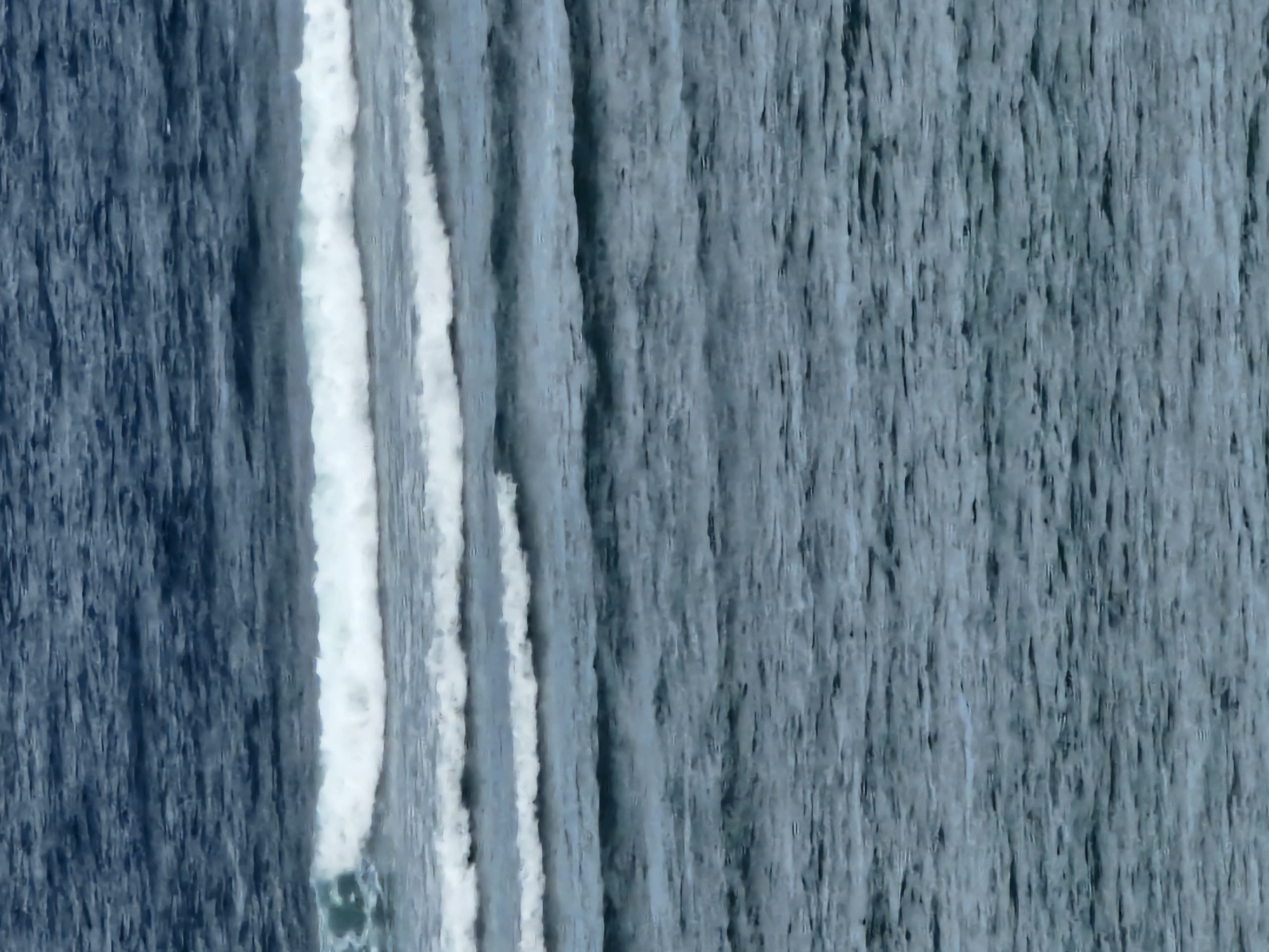 The image is a close-up, high-angle view of choppy ocean water with several waves. The water is a deep blue-grey color, with white foam cresting on the waves. The waves are generally parallel to each other, moving from left to right across the frame, with smaller ripples and disturbances visible throughout the water's surface. There are no visible people, man-made objects, or landmasses. The scene is solely focused on the movement and texture of the ocean.