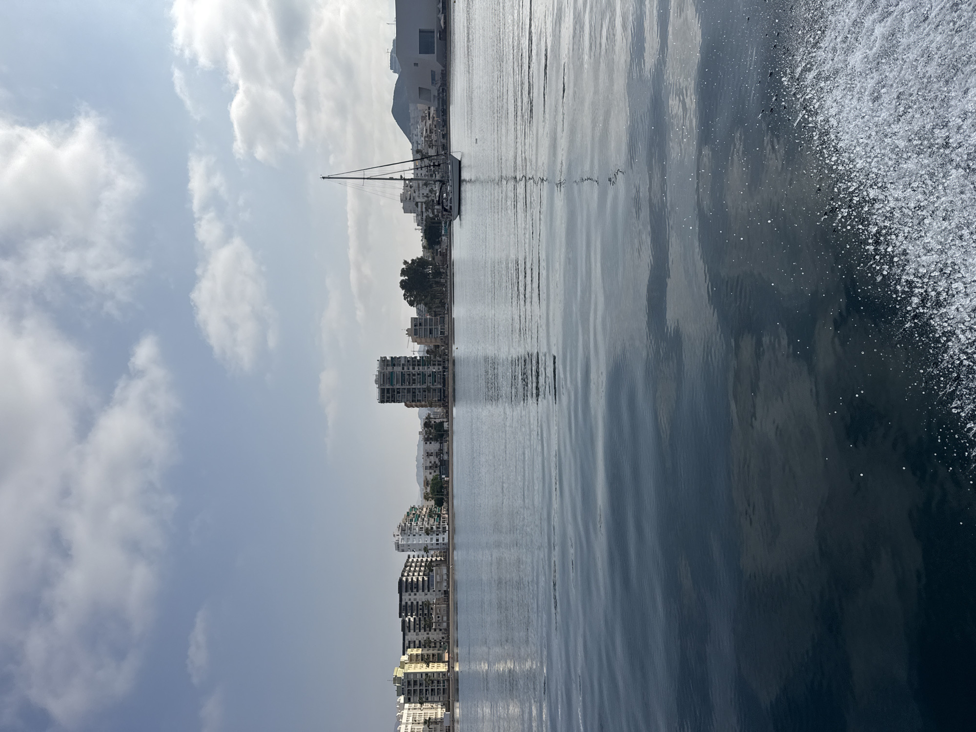 The image depicts a calm bay with a modern cityscape on the horizon. Several multi-story buildings, some with balconies and glass facades, line the waterfront. A prominent sailboat with a dark mast and hull is anchored in the middle of the bay, its reflection rippling on the water's surface. The sky above is a pale blue, dotted with scattered white clouds. The foreground features the choppy wake of a boat, with white spray and bubbles indicative of movement. The overall scene suggests a tranquil coastal environment in Águilas, Spain, likely a harbor or marina area.