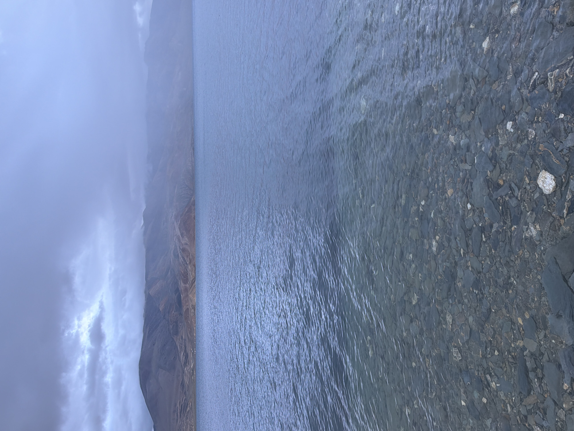 The image depicts a serene, natural landscape scene on a cloudy day. The foreground is dominated by the clear, shallow waters of a lake, revealing a rocky, pebble-strewn bottom. Ripples gently disturb the surface of the water. In the midground, the expanse of the lake stretches towards the horizon, its surface reflecting the muted colors of the sky. The background features a range of rugged, barren mountains under a thick, overcast sky. The mountains exhibit varied tones of brown and grey, with some areas softened by mist or clouds clinging to their peaks. A small cluster of structures, possibly dwellings or buildings, is visible nestled at the base of the mountains along the shoreline, suggesting a settlement. The overall atmosphere is one of tranquility and natural beauty, with the cloudy sky lending a dramatic and subdued mood to the scene. This scene is consistent with the location of Karzok, India, a village situated on the shores of Tso Moriri lake in the Ladakh region.